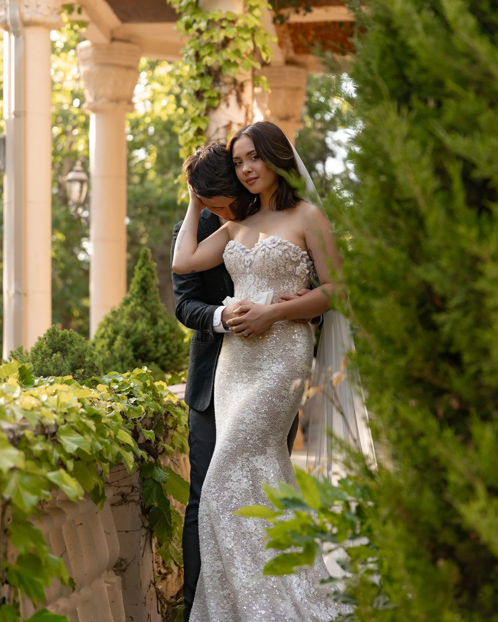 A bride and groom embracing outdoors, surrounded by greenery and old columns, during a wedding photoshoot in late afternoon sunlight.