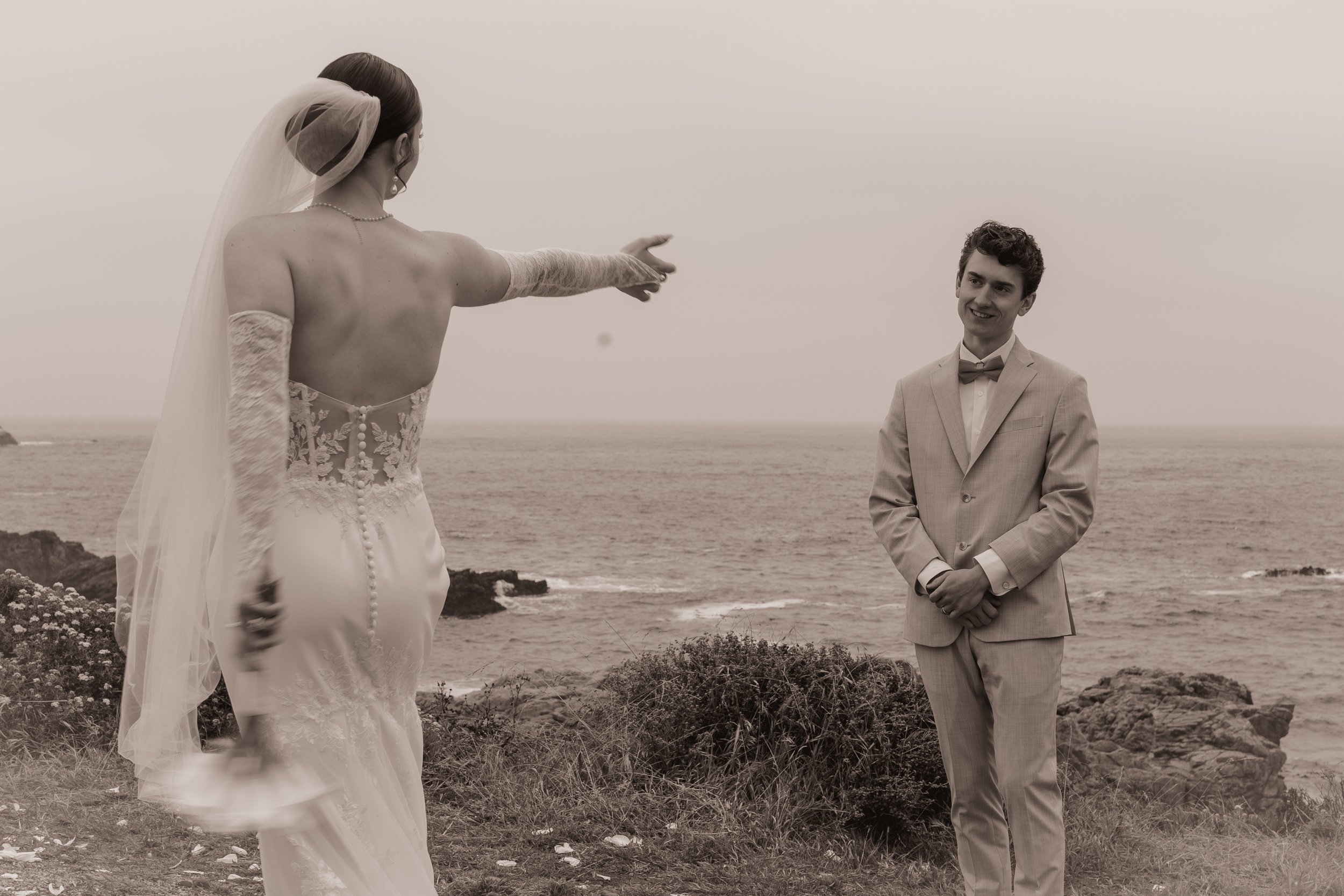 A bride pointing at a groom on a beach during sunset, with the ocean in the background.