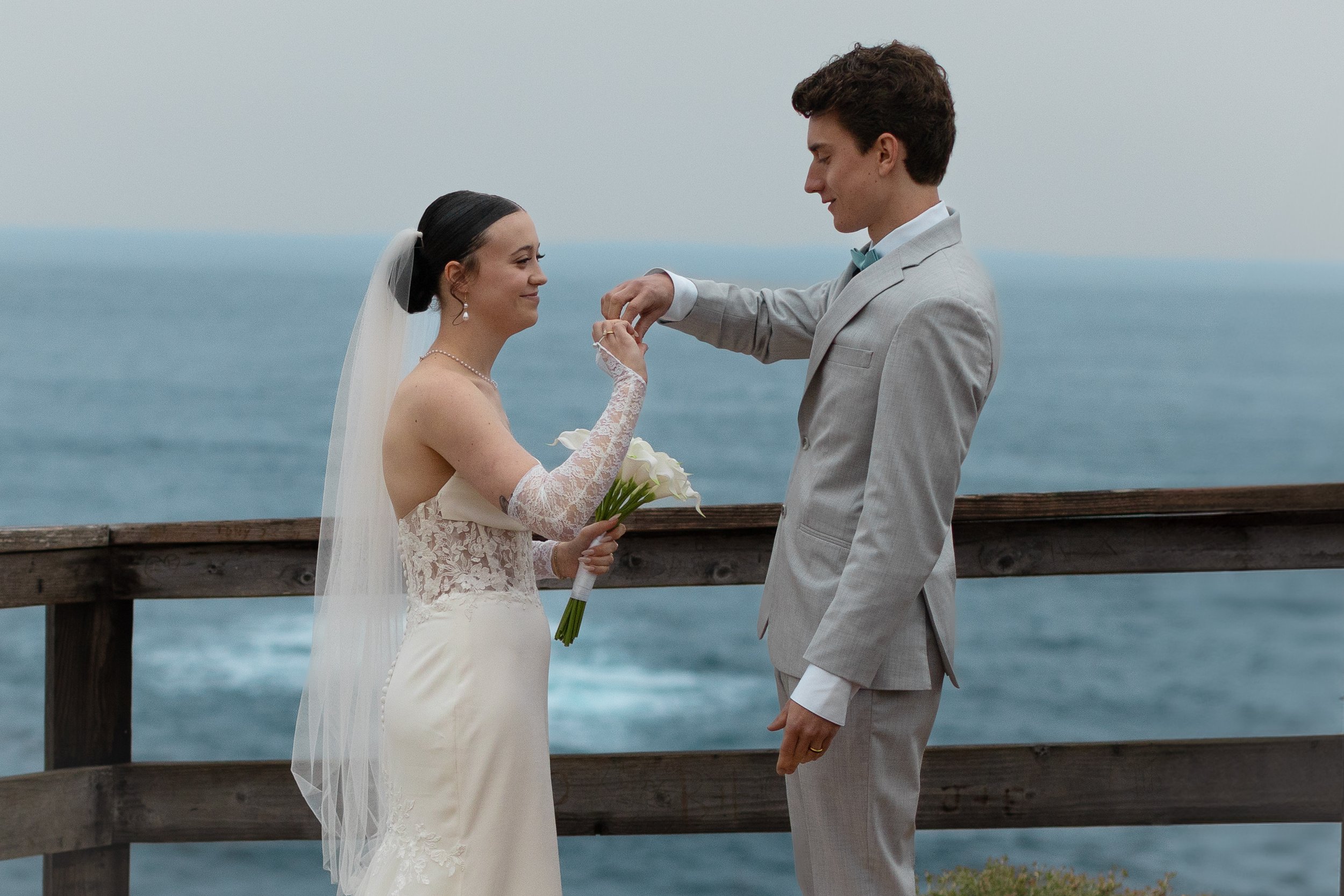 A bride and groom exchange rings on a cliff overlooking the ocean during their wedding ceremony.