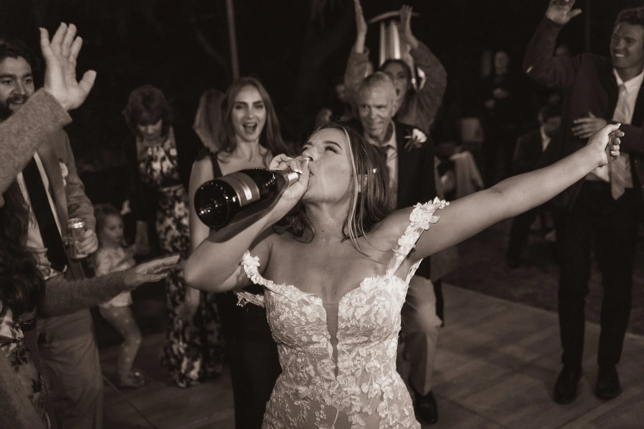 A woman in a wedding dress is drinking from a bottle of champagne at a celebration with friends and family dancing and celebrating around her.