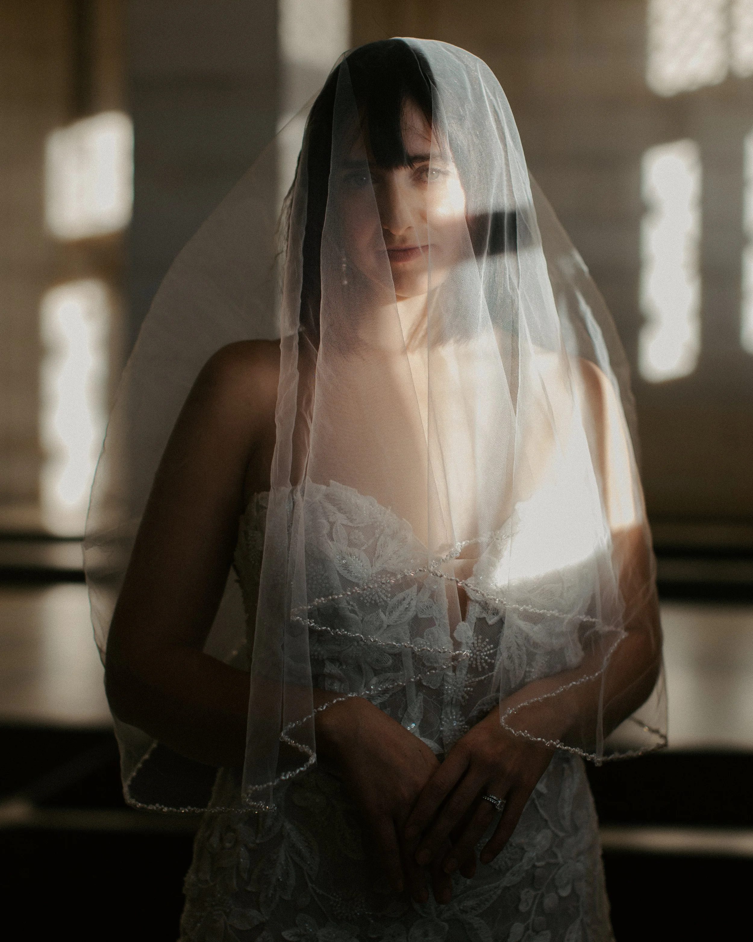 Woman in wedding dress with veil standing indoors, soft natural light illuminating her face, hands clasped in front, rings visible.
