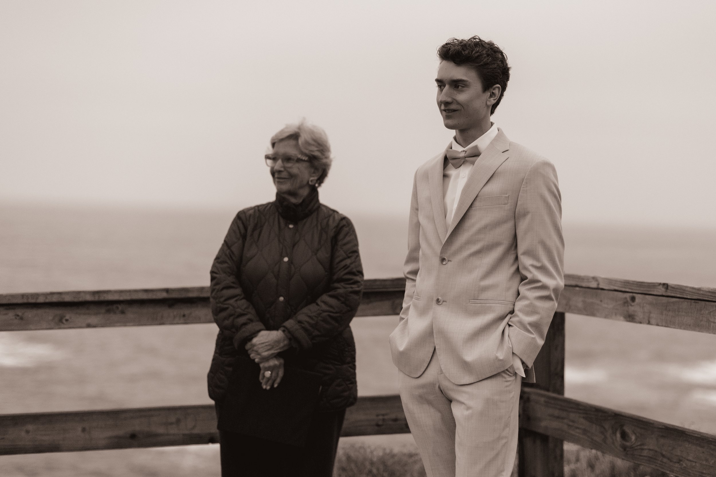 A young man in a light-colored suit and bow tie standing next to an older woman in a dark quilted jacket, both looking off into the distance on a wooden pier with a blurred ocean background.