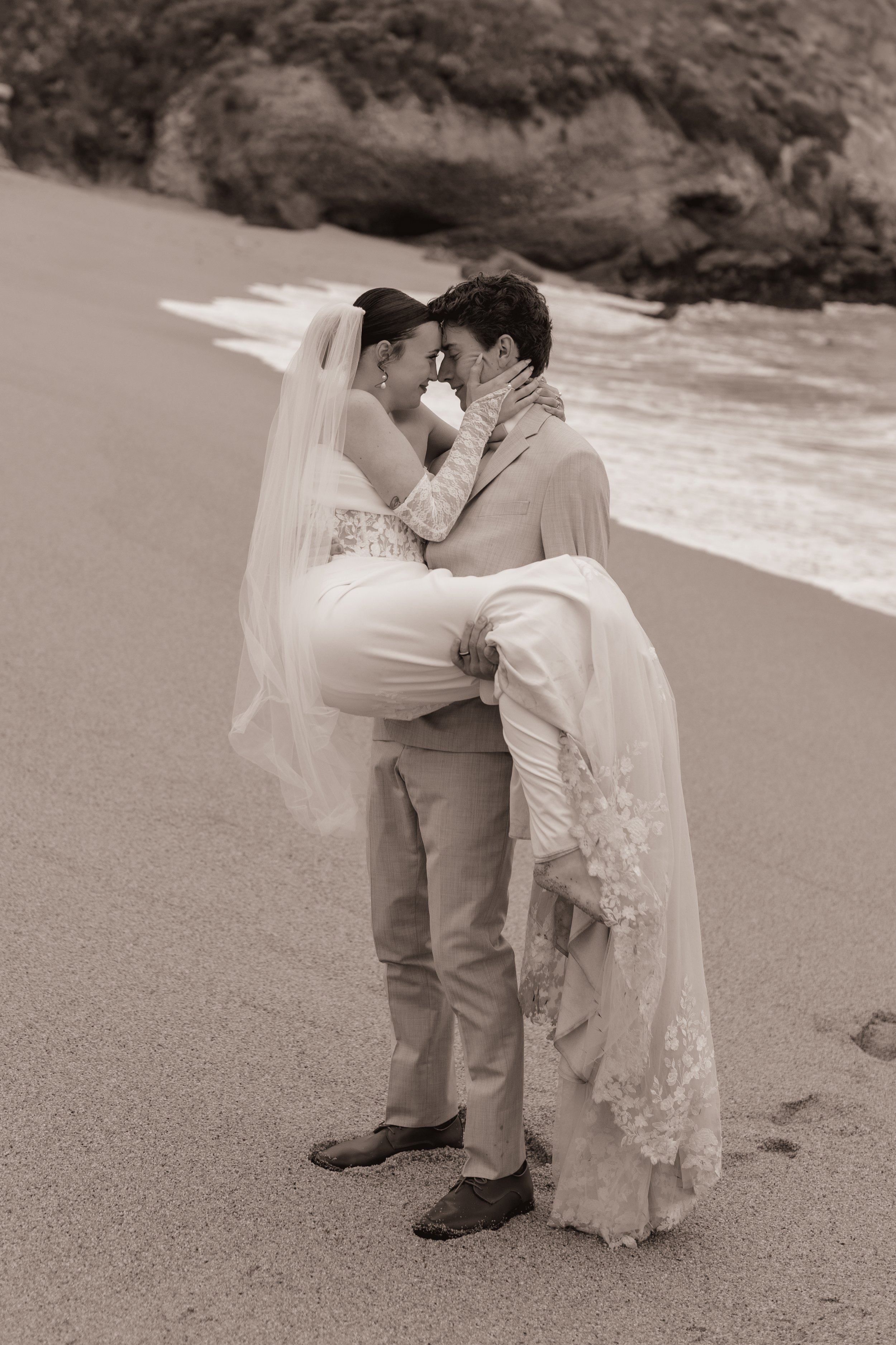 A newlywed couple on a sandy beach, with one person in a wedding dress being carried by the other, as they touch foreheads and smile at each other.