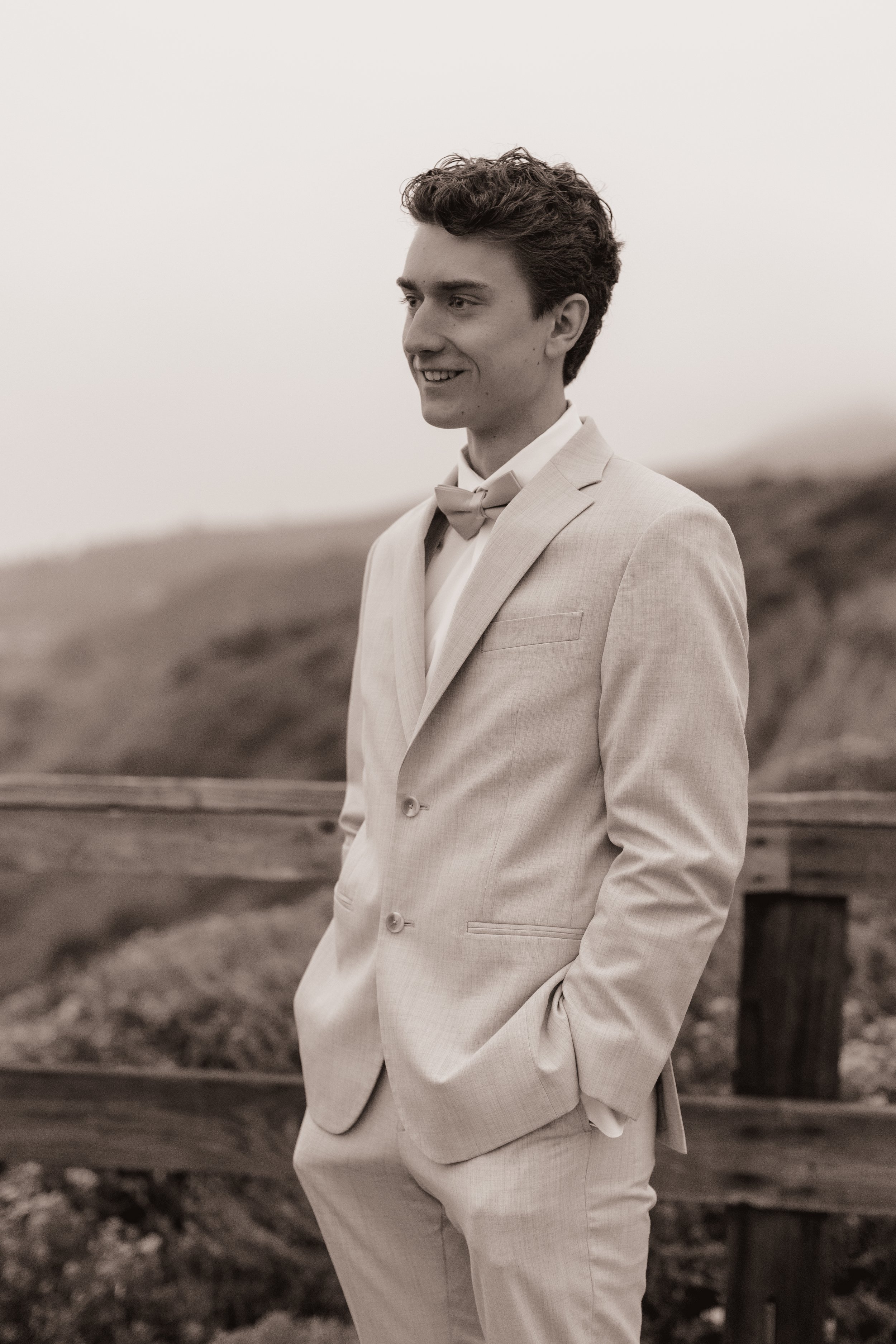 A young man in a light-colored suit and bow tie, standing outdoors on a cloudy day, smiling with hands in pockets.