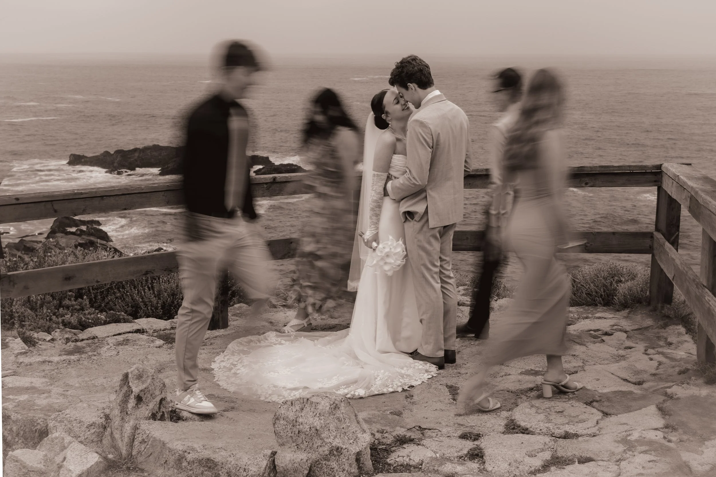 A bride and groom share a kiss on a rocky coastline during their wedding, with blurred guests walking around them, in black and white.