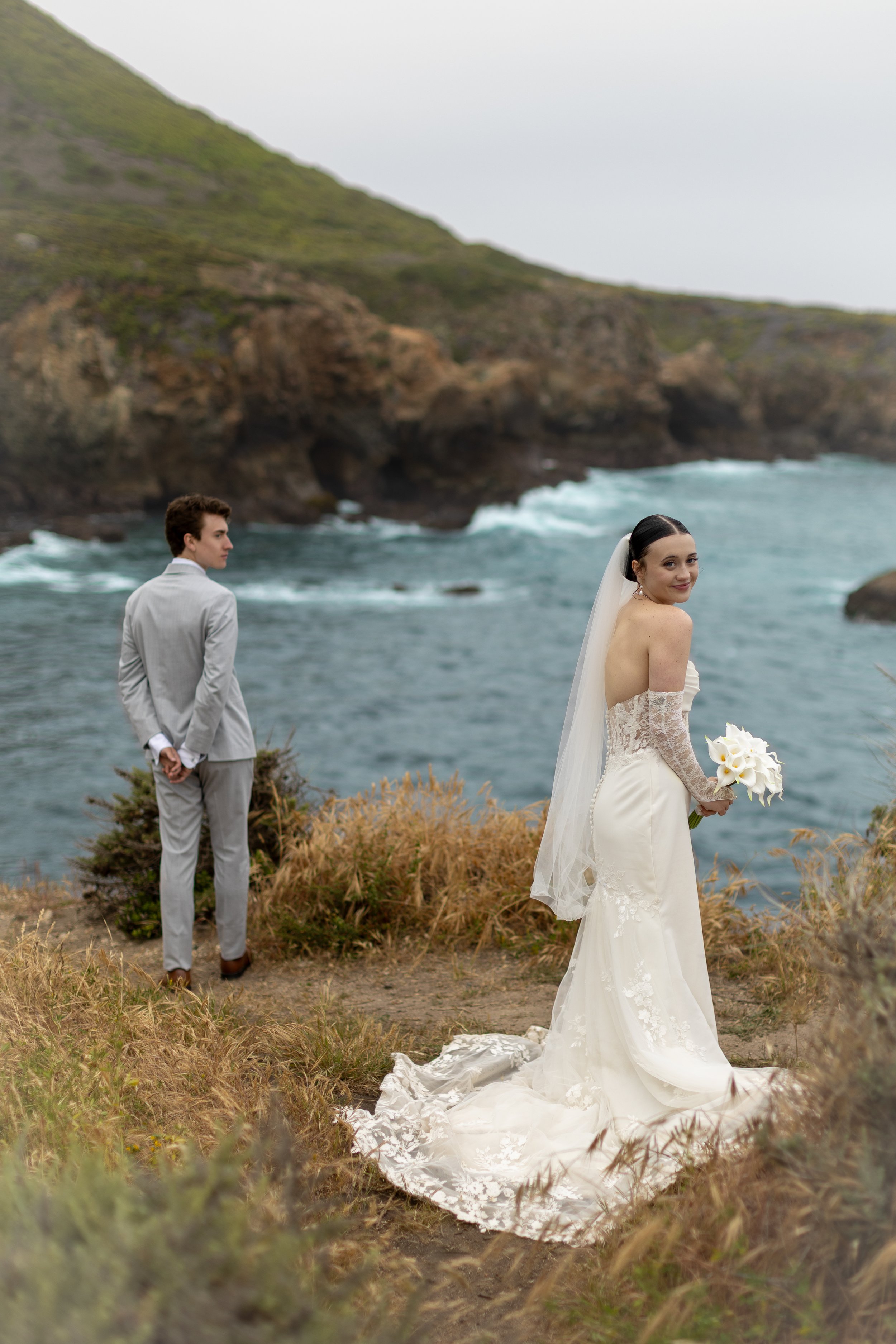 A bride in a white wedding dress holding flowers looks back at a groom in a grey suit standing near the edge of a rocky coastline, with ocean waves and green hills in the background.