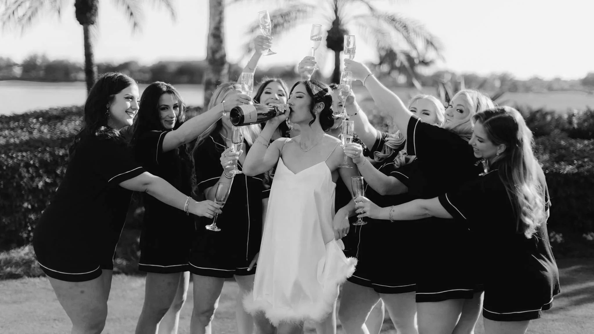 Group of women celebrating, many holding champagne glasses and a bottle, outdoors with palm trees in the background, black and white photo.