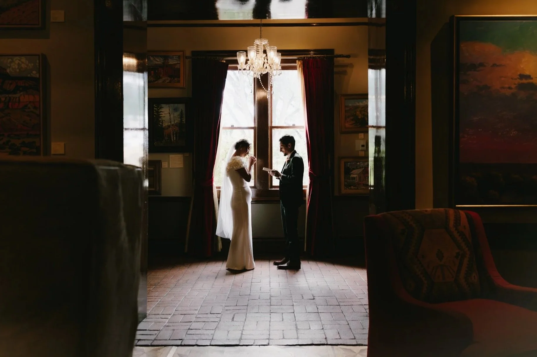 A bride and groom standing in front of a large window with light streaming in, holding hands and exchanging vows during a wedding ceremony inside an art-filled room with a chandelier overhead at La Posada Santa Fe Spa and Hotel. 