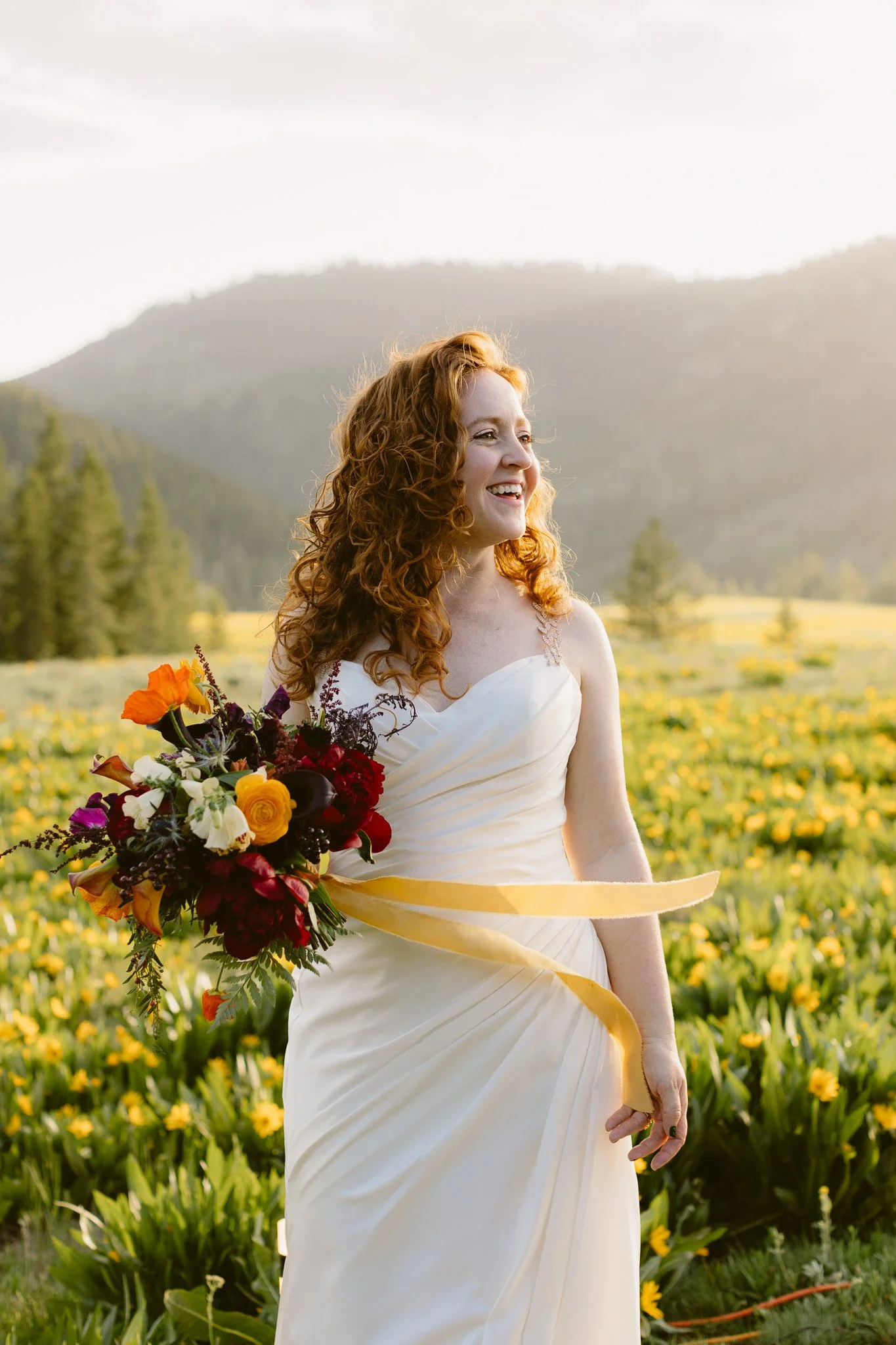 A woman with curly red hair wearing a white dress, holding a colorful bouquet of flowers, standing in a field of yellow flowers with mountains in the background.