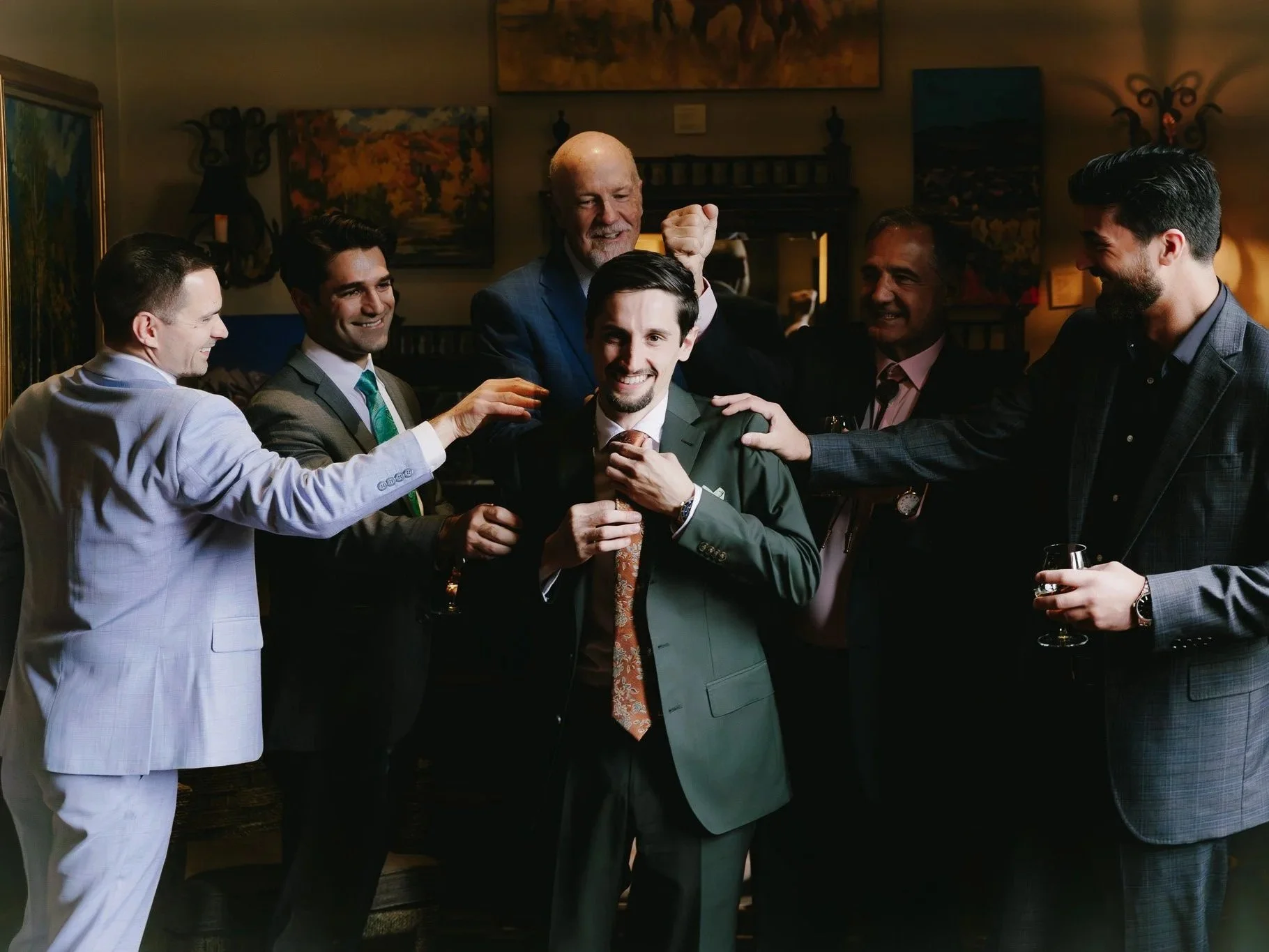 A group of six men in suits smiling and celebrating together, with one man being the center of attention, in a warm, indoor setting