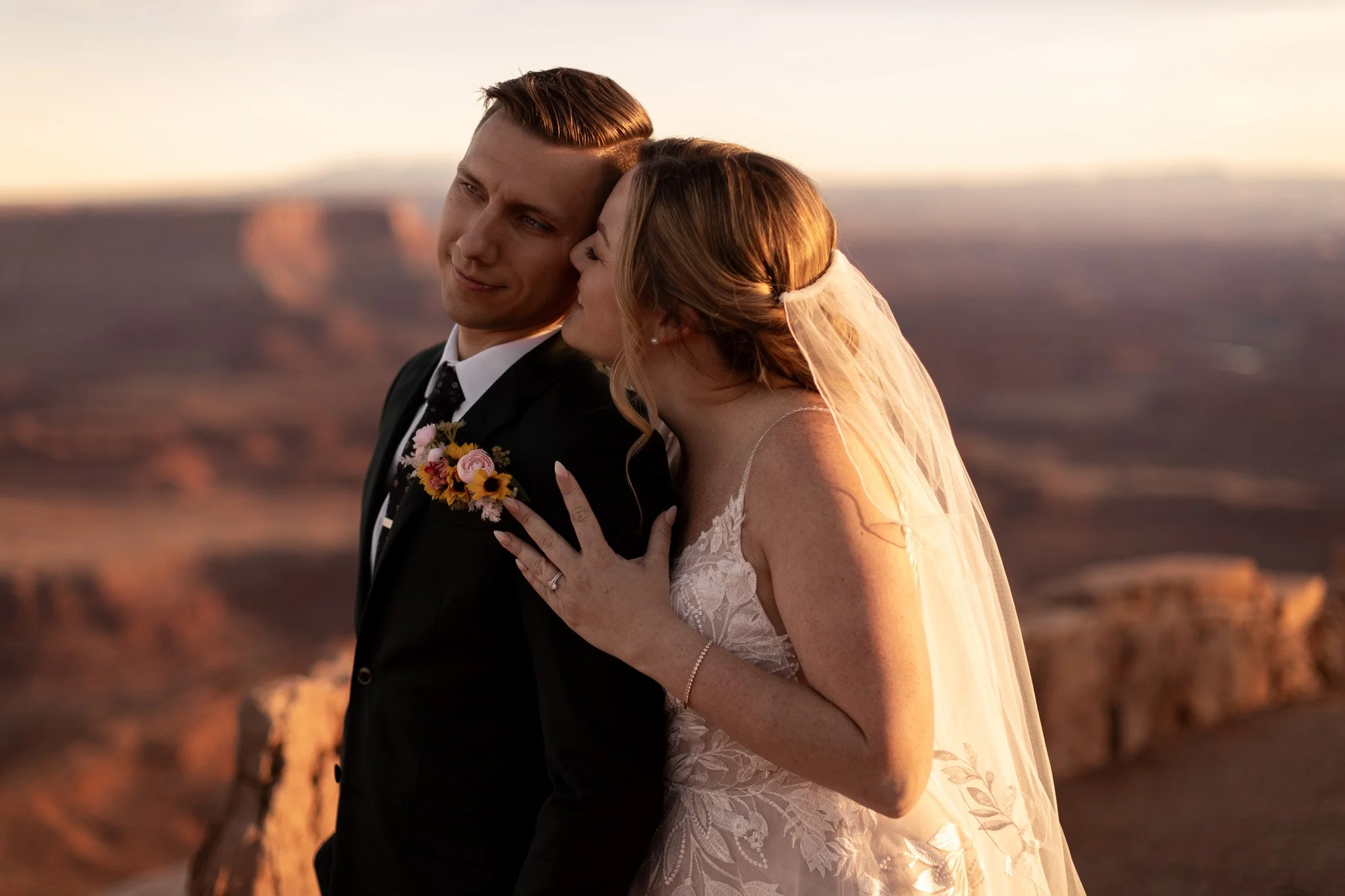 A bride and groom on their wedding day, with the bride kissing the groom's cheek at deadhorse state park sunset with a canyon landscape in the background.