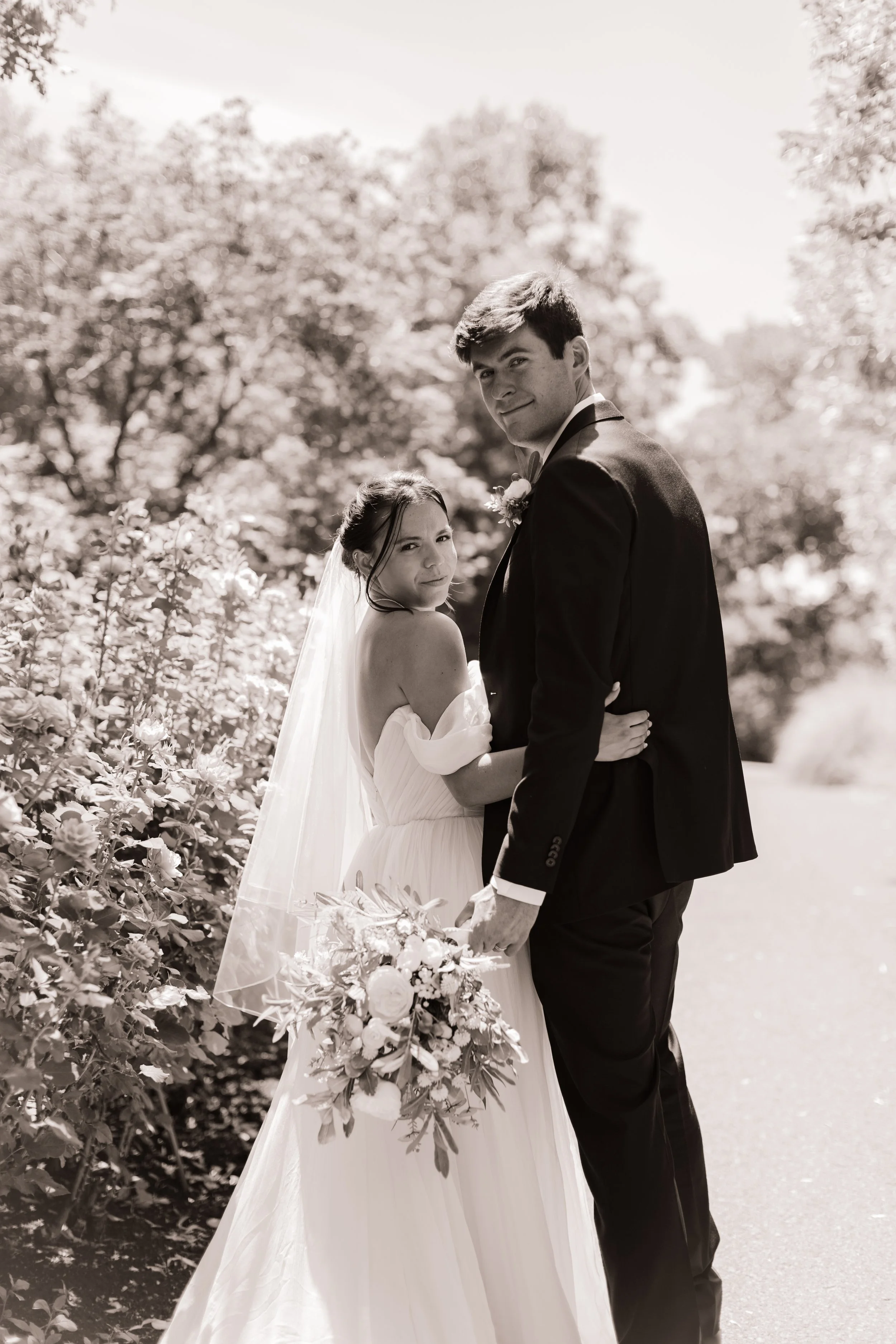 Black and white photo of a bride and groom standing outdoors, holding a bouquet of flowers, with trees in the background.