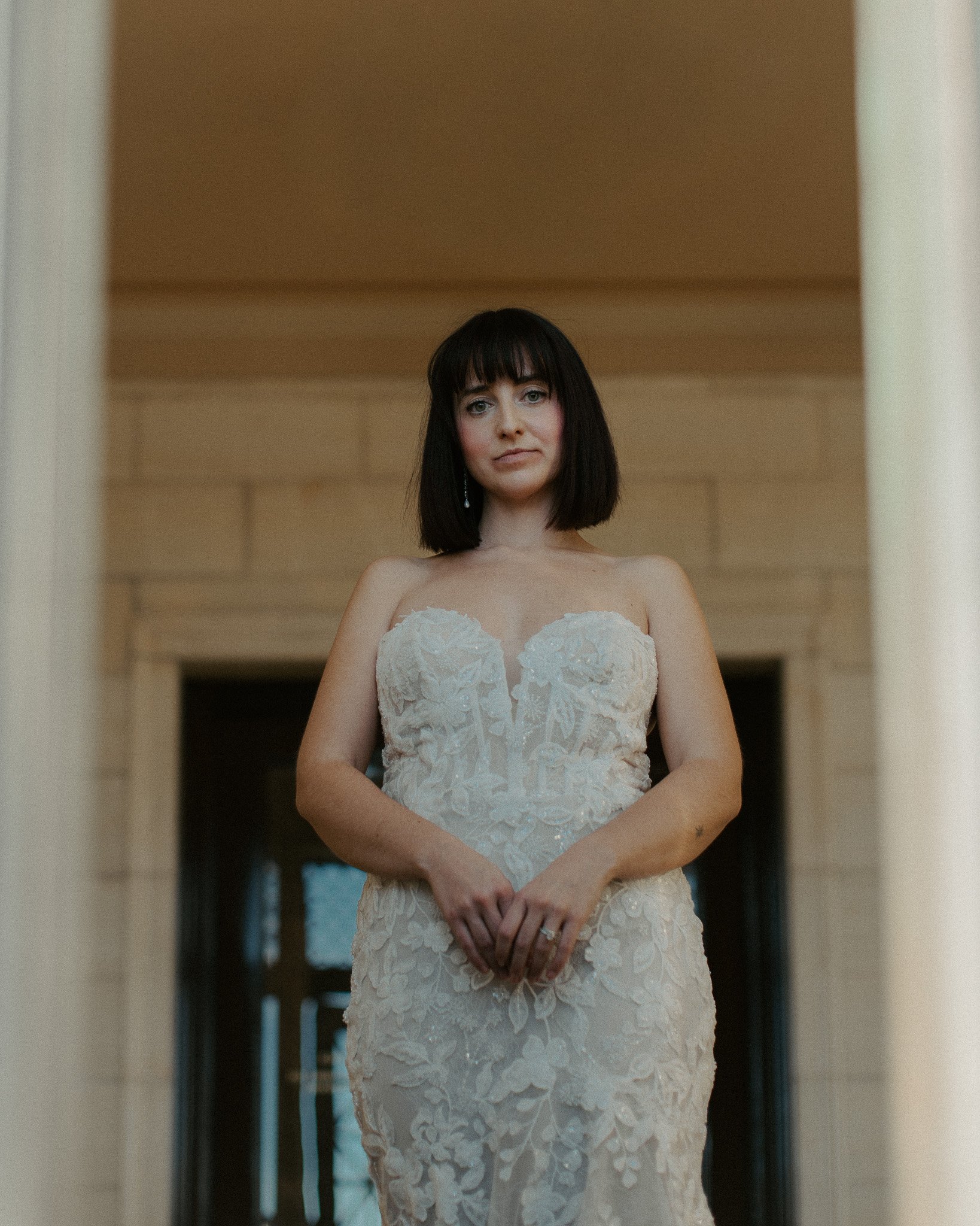 A woman in a wedding dress standing indoors, looking at the camera with a neutral expression.