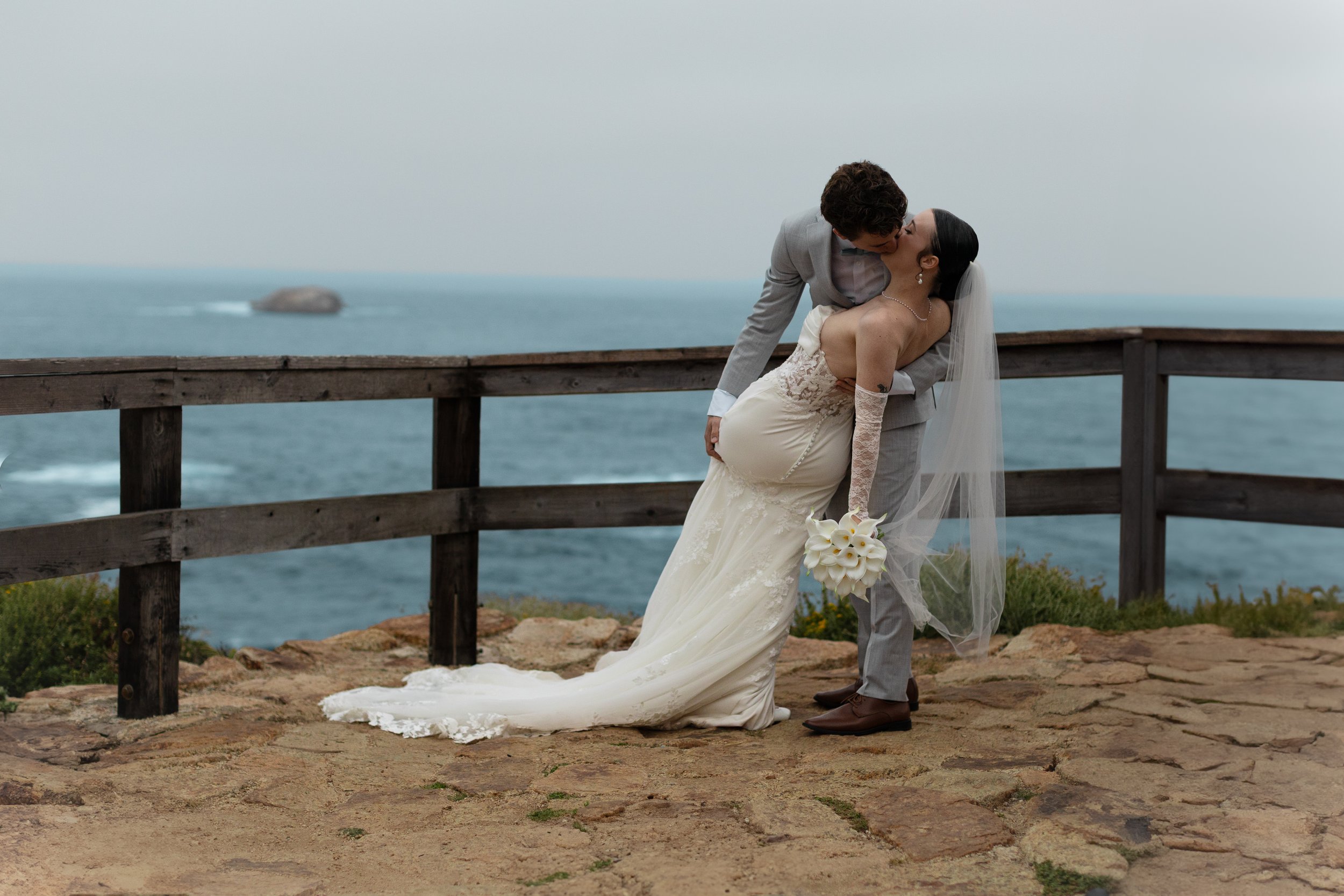 A bride and groom sharing a romantic kiss by the ocean on a rocky path, with a wooden railing and a distant rock in the water.