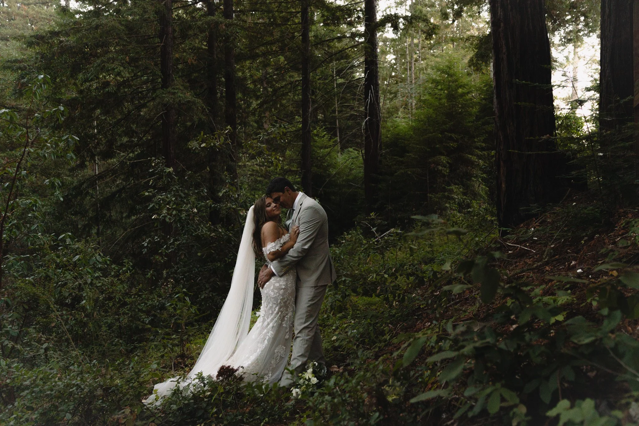 A newlywed couple embracing in a forest, the bride in a white wedding gown with lace details and long veil, the groom in a light gray suit, surrounded by tall trees and greenery.