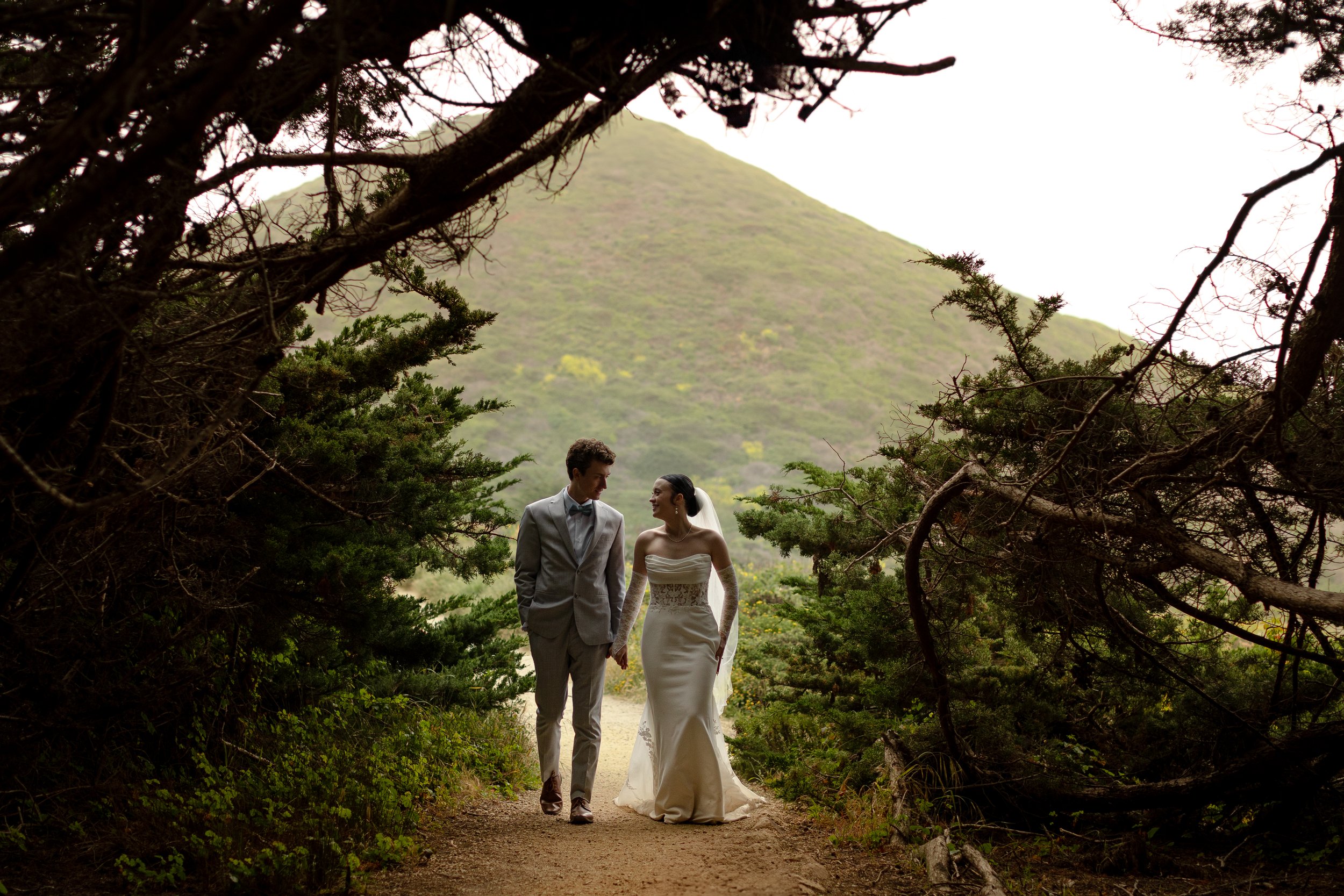 A bride and groom walking hand in hand on a dirt path through a forested area with mountains in the background.
