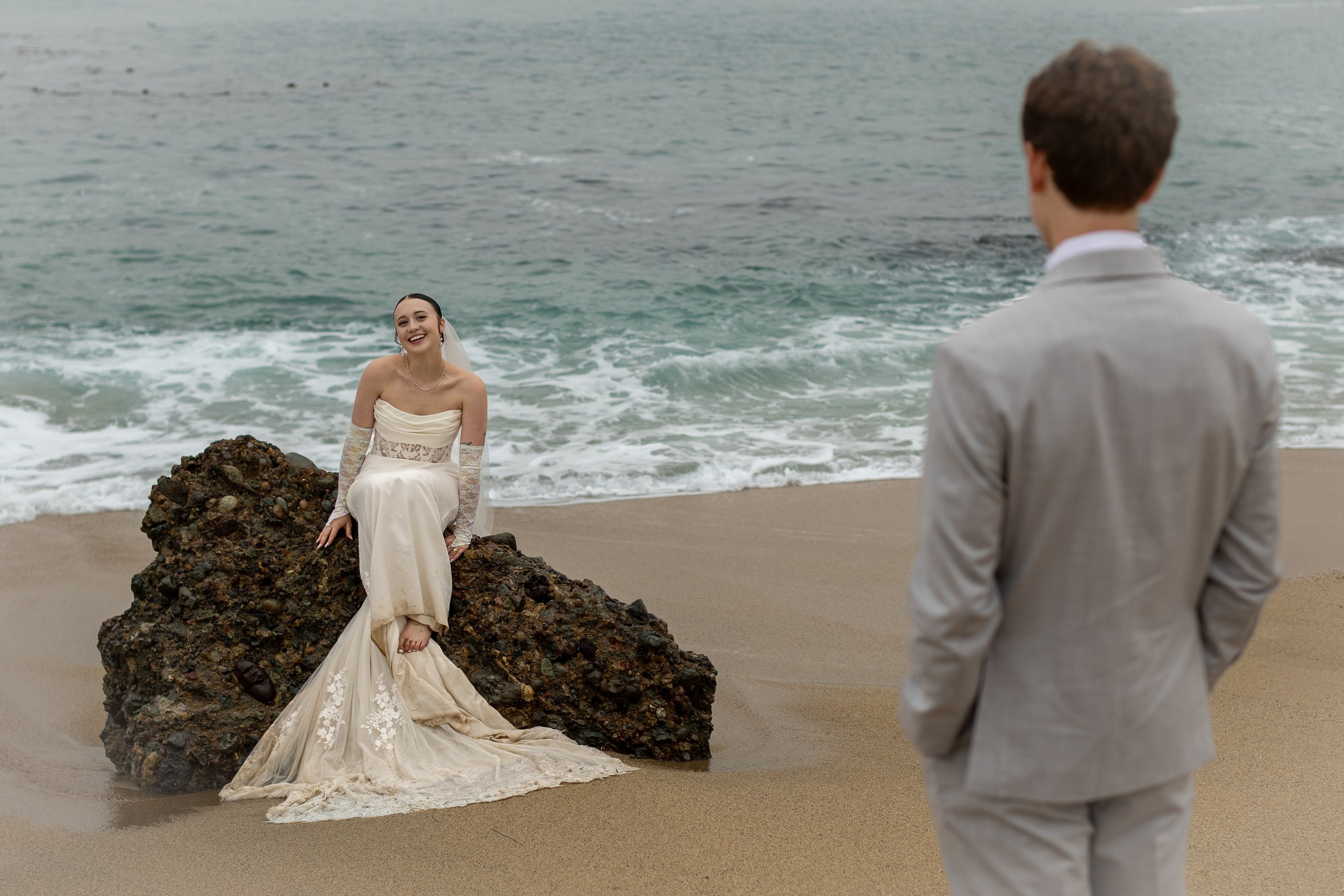 A woman in a wedding dress sitting on a rock at the beach, smiling, with a man in a gray suit watching her.