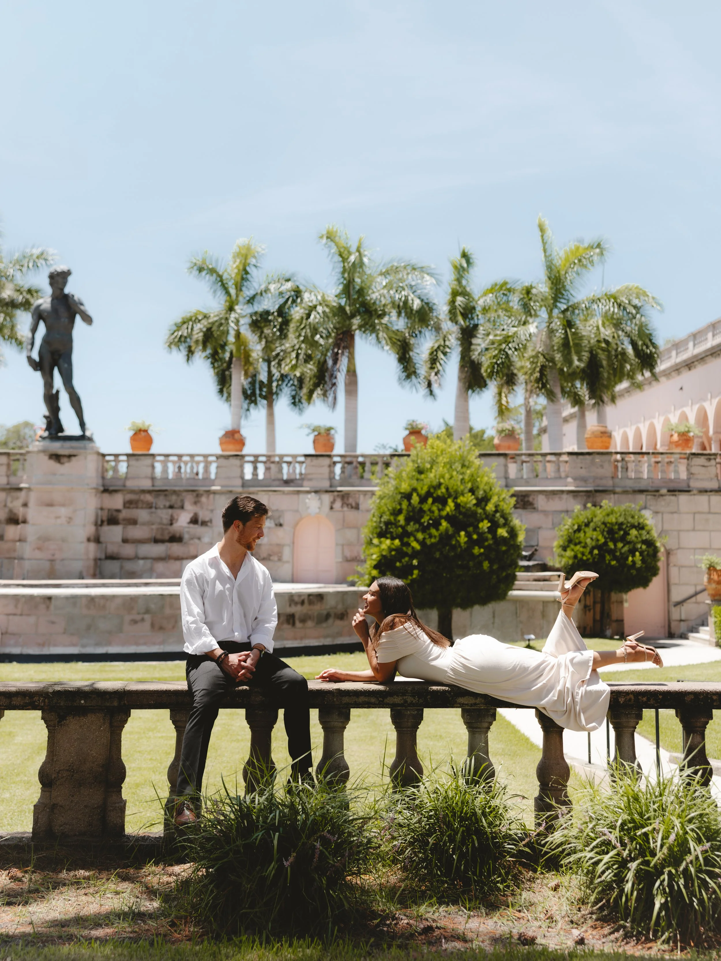 A man in a white shirt and black pants kneels and looks at a woman in a white dress lying on a stone ledge in a garden with trees and a castle-like building in the background.