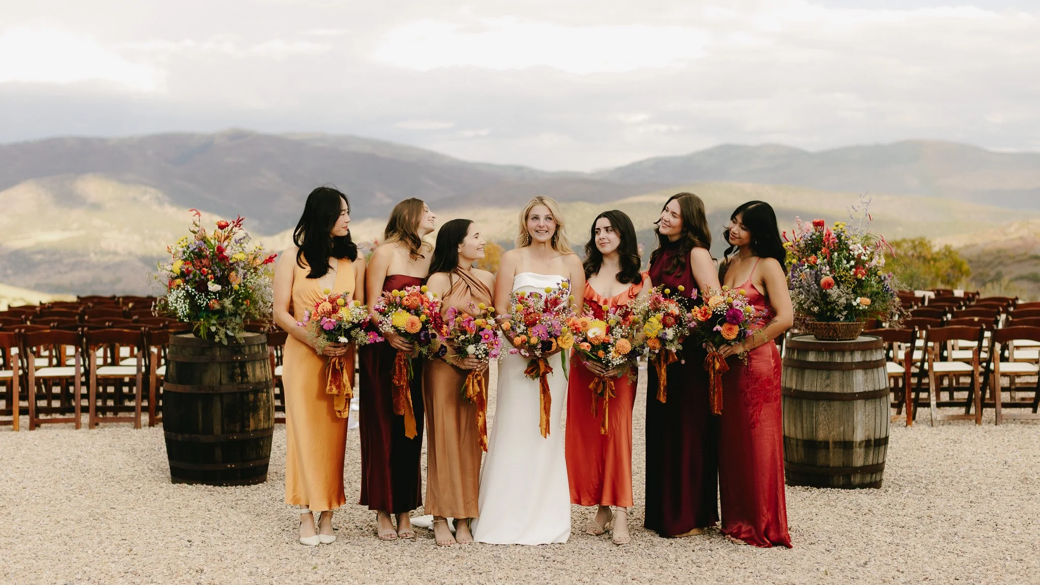A group of seven women in colorful dresses standing in a row outdoors, holding large bouquets of flowers, with open sky and rolling hills in the background.