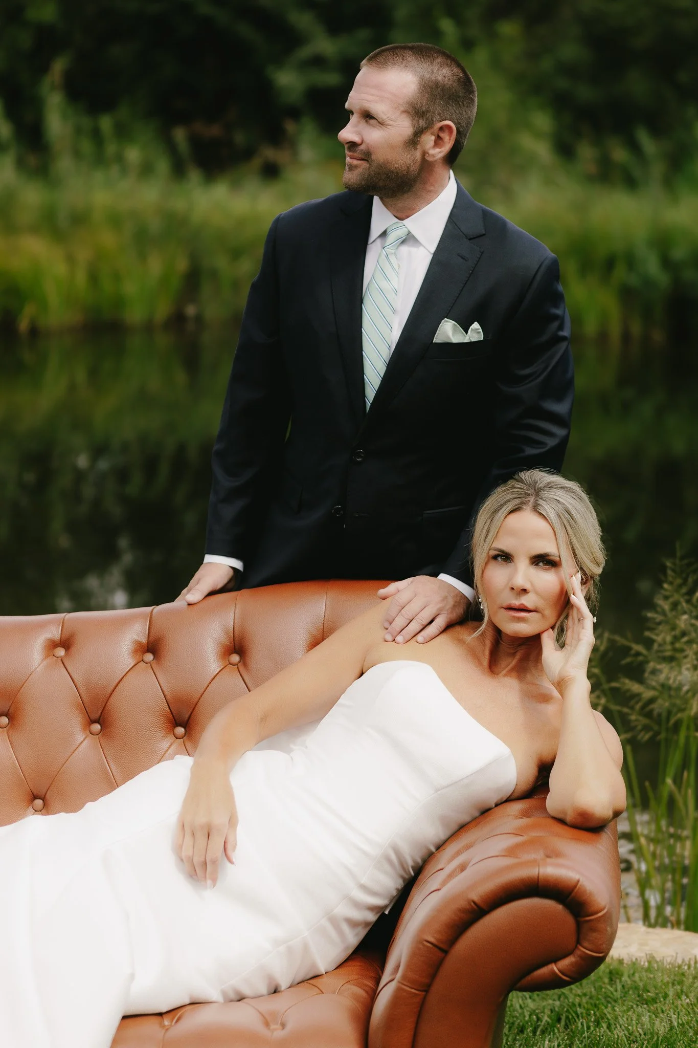 A man in a suit standing behind a woman lying on a brown leather tufted sofa outdoors near a pond with green foliage in the background.