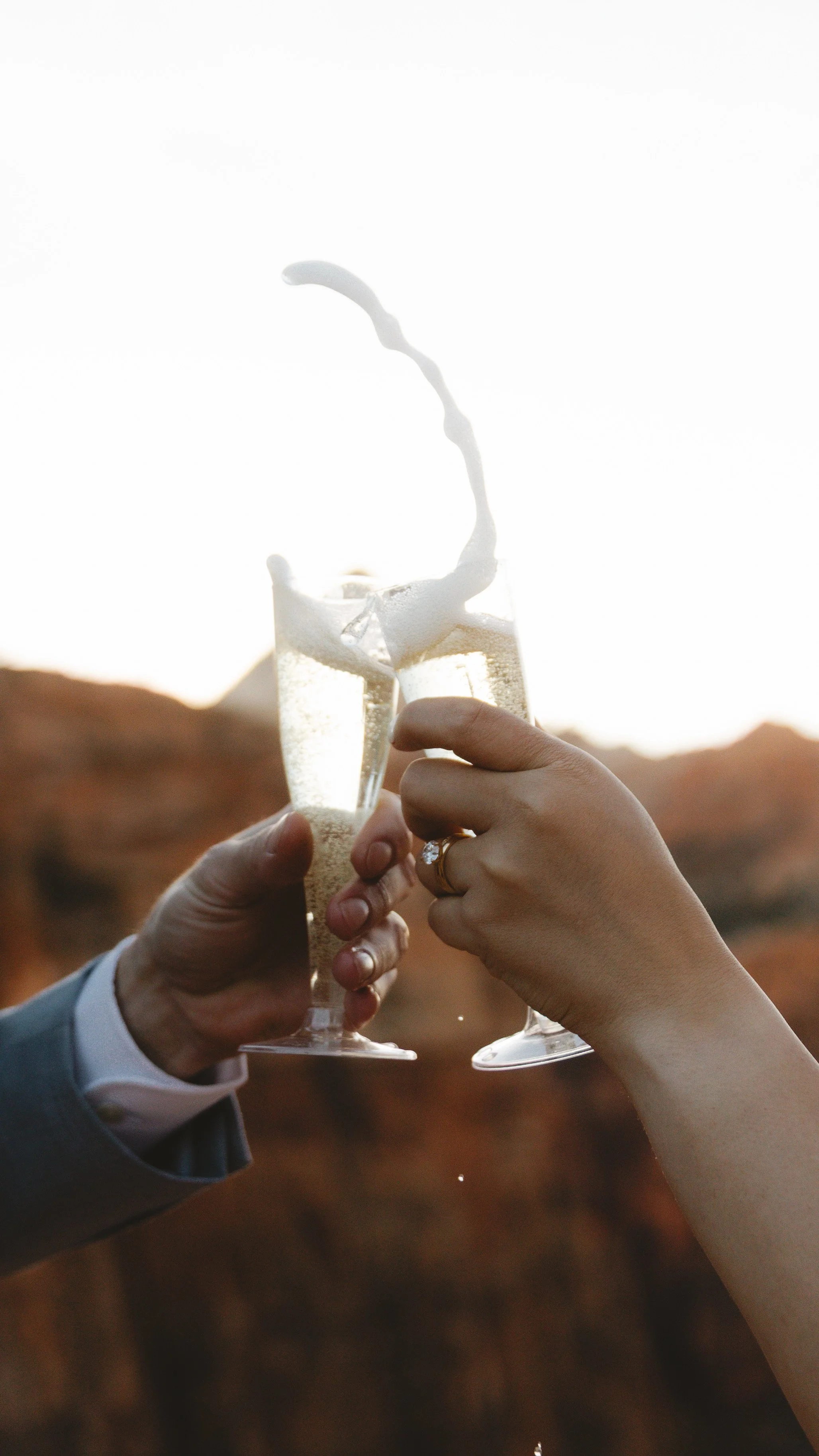 Two people holding champagne glasses clinking, with a splash of champagne and a blurry outdoor landscape in the background.