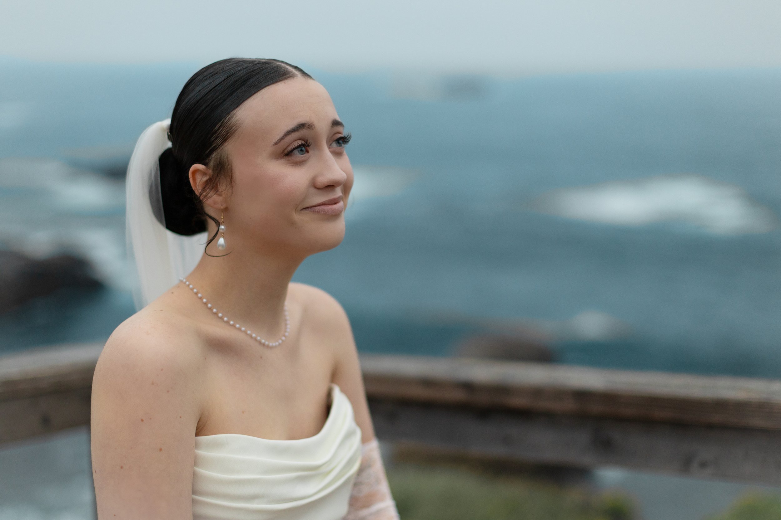 A young woman in a wedding dress standing outdoors by the water, with a serene expression and a gentle smile.