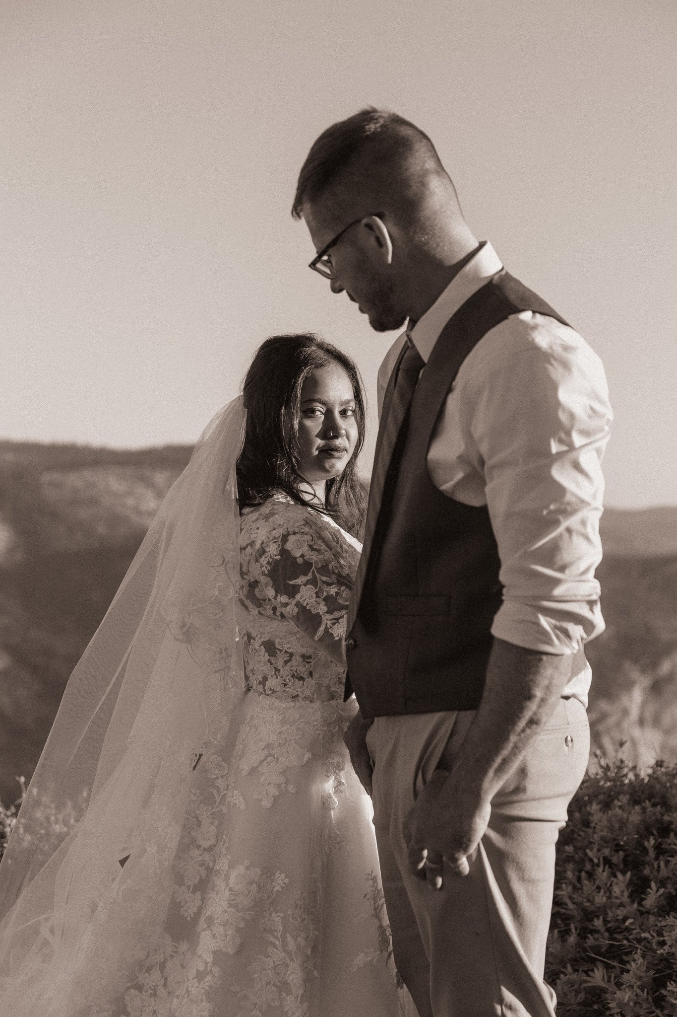Sepia-toned photo of a bride and groom in wedding attire outdoors; the bride wears a lace dress with a veil, and the groom wears a vest and tie.