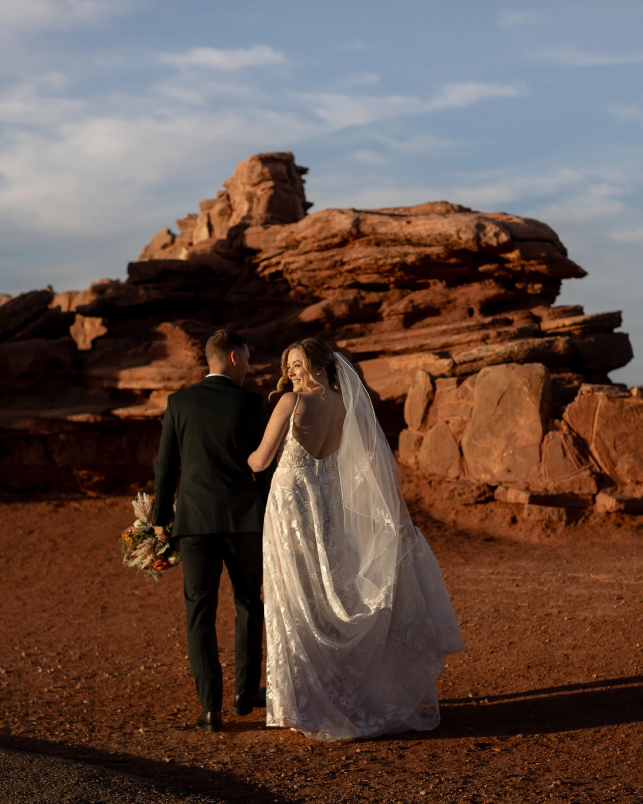 A bride and groom walking hand in hand in a desert landscape with red rocks, during sunset, with the bride smiling at the groom.
