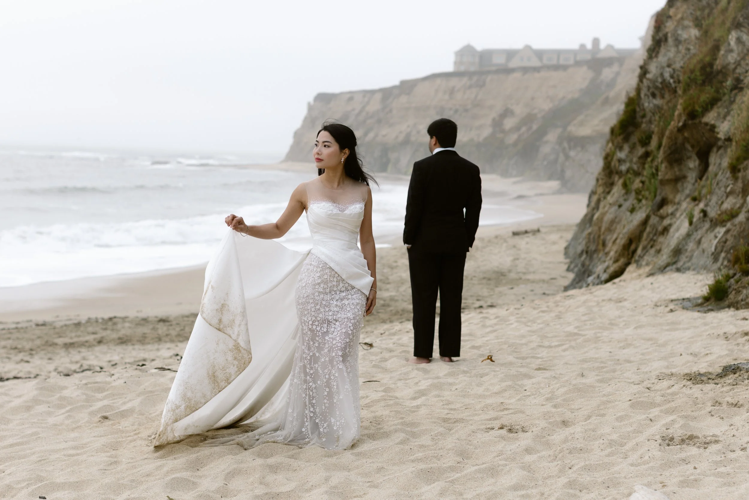 A woman in a white wedding dress holding the train standing on a sandy beach, with a man in a black suit facing away in the background, along a rocky coastline with cliffs and buildings in the distance.