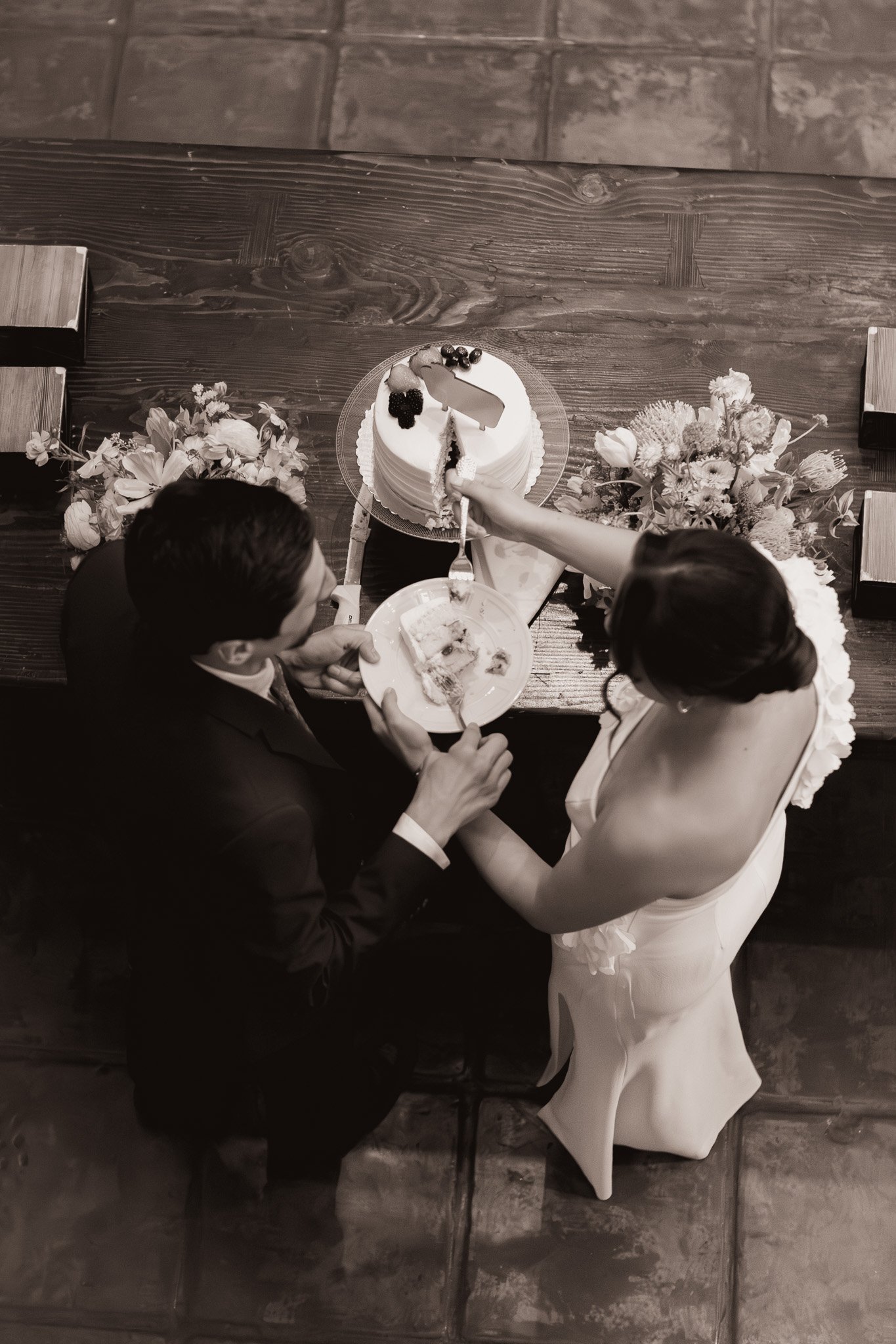 A bride and groom sharing a wedding cake and cake at their wedding reception, seen from above, with flowers on a wooden table.