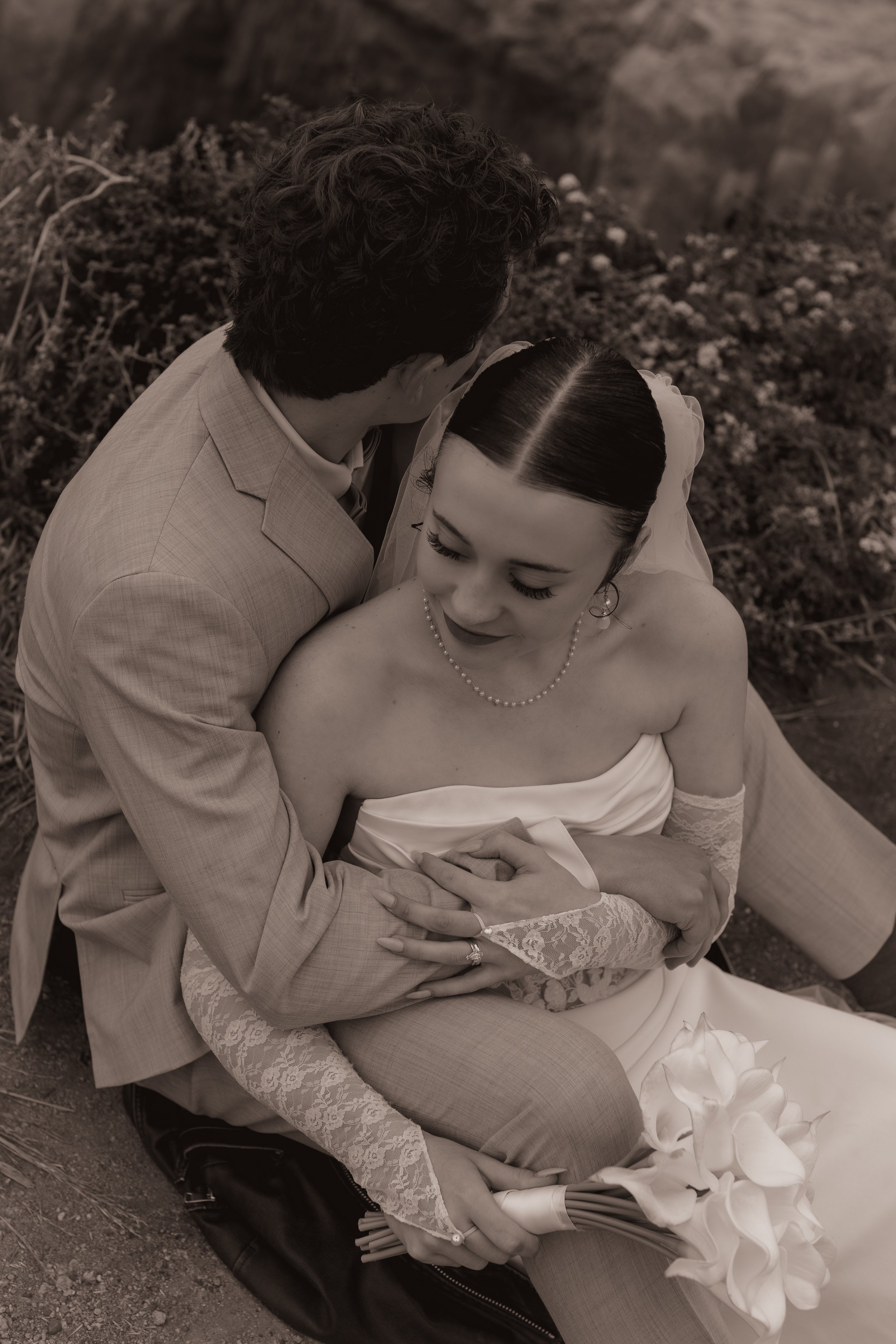 A bride sitting on the ground holding a bouquet of calla lilies, embraced by a man in a light-colored suit, with her eyes closed and a gentle smile.