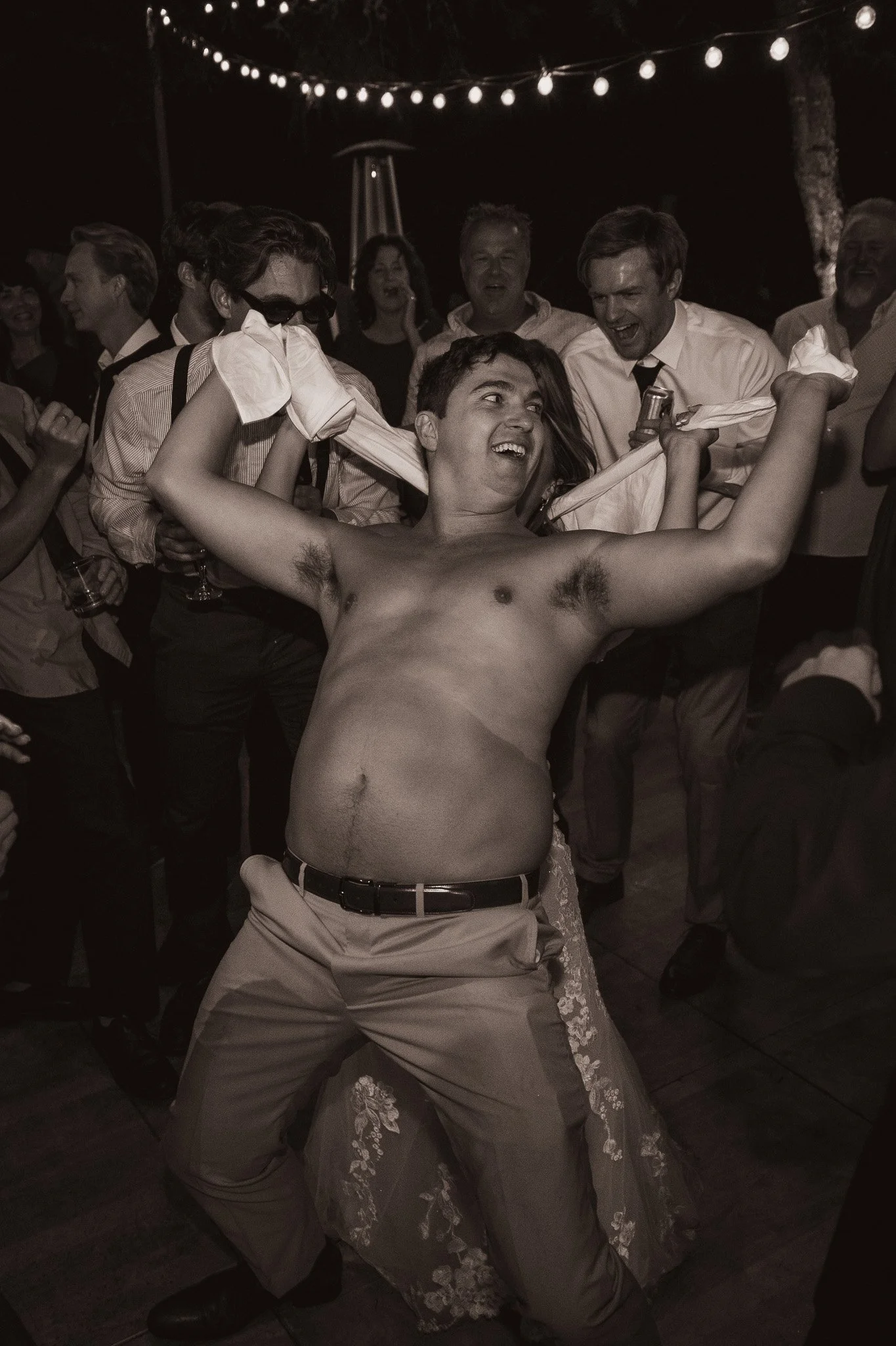 A shirtless man with a large beer gut dance enthusiastically at a party surrounded by smiling guests. String lights hang above.