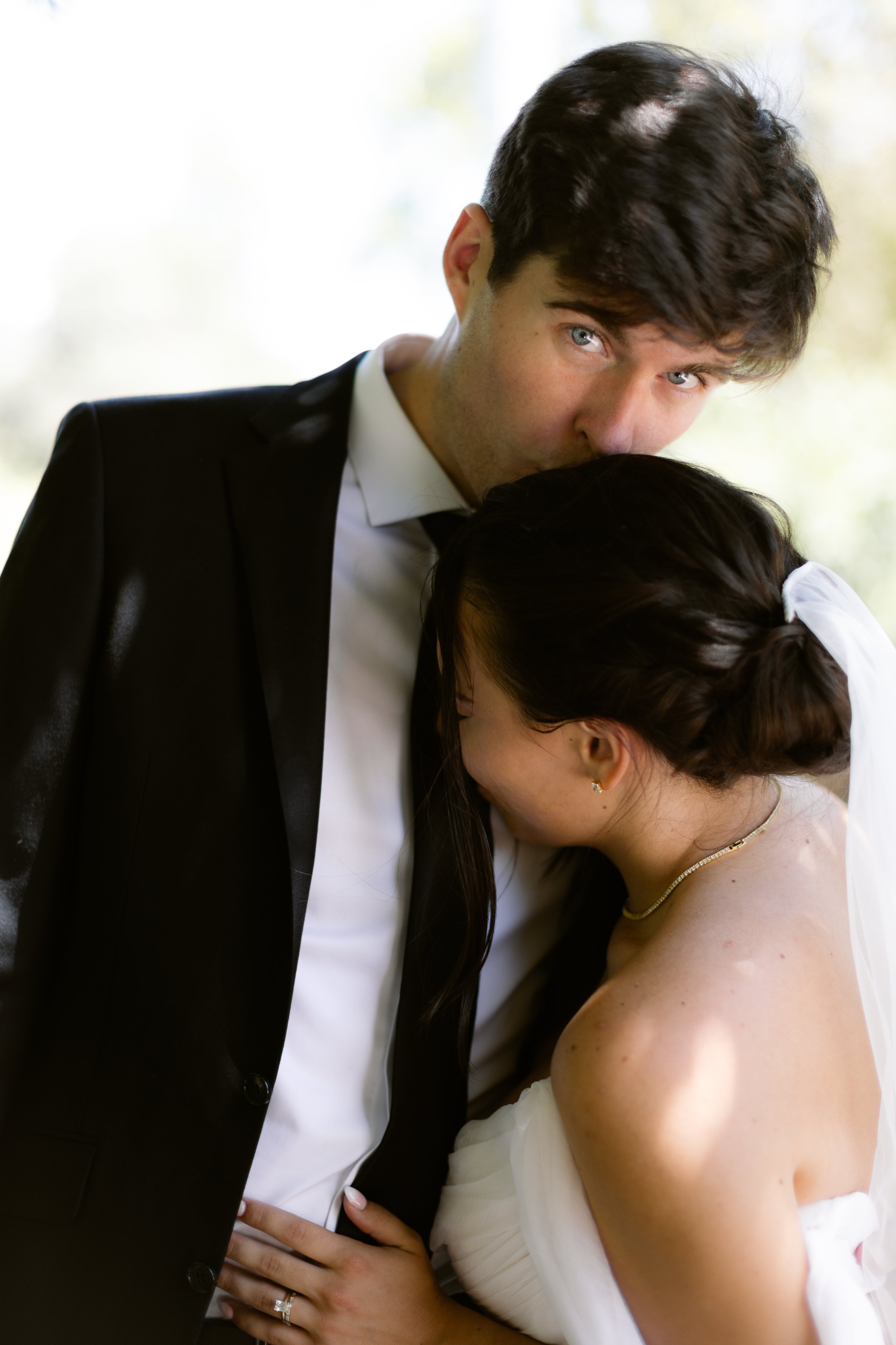 A wedding couple is sharing an intimate moment, with the groom kissing the bride's forehead as she rests her head on his chest.