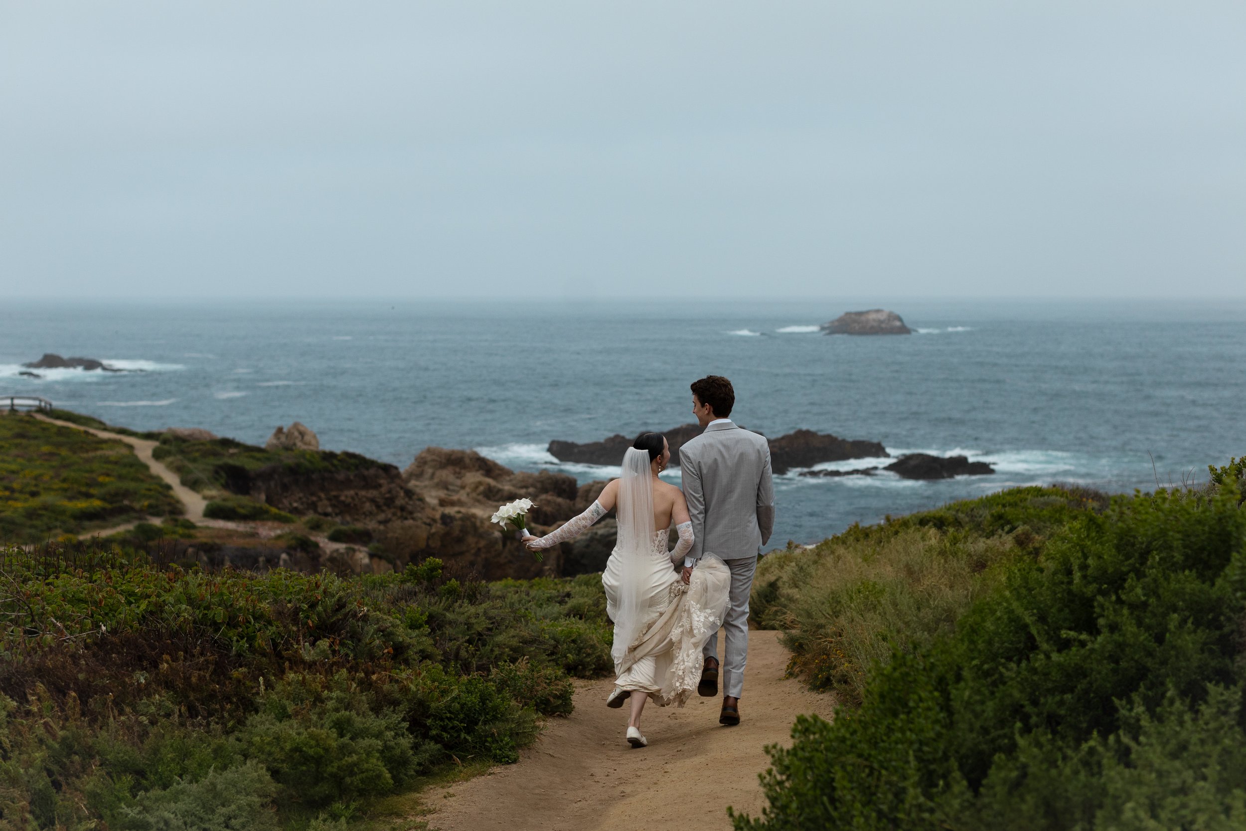 Bride and groom walking along a coastal dirt path, holding hands, with the ocean and rocky islands in the background.