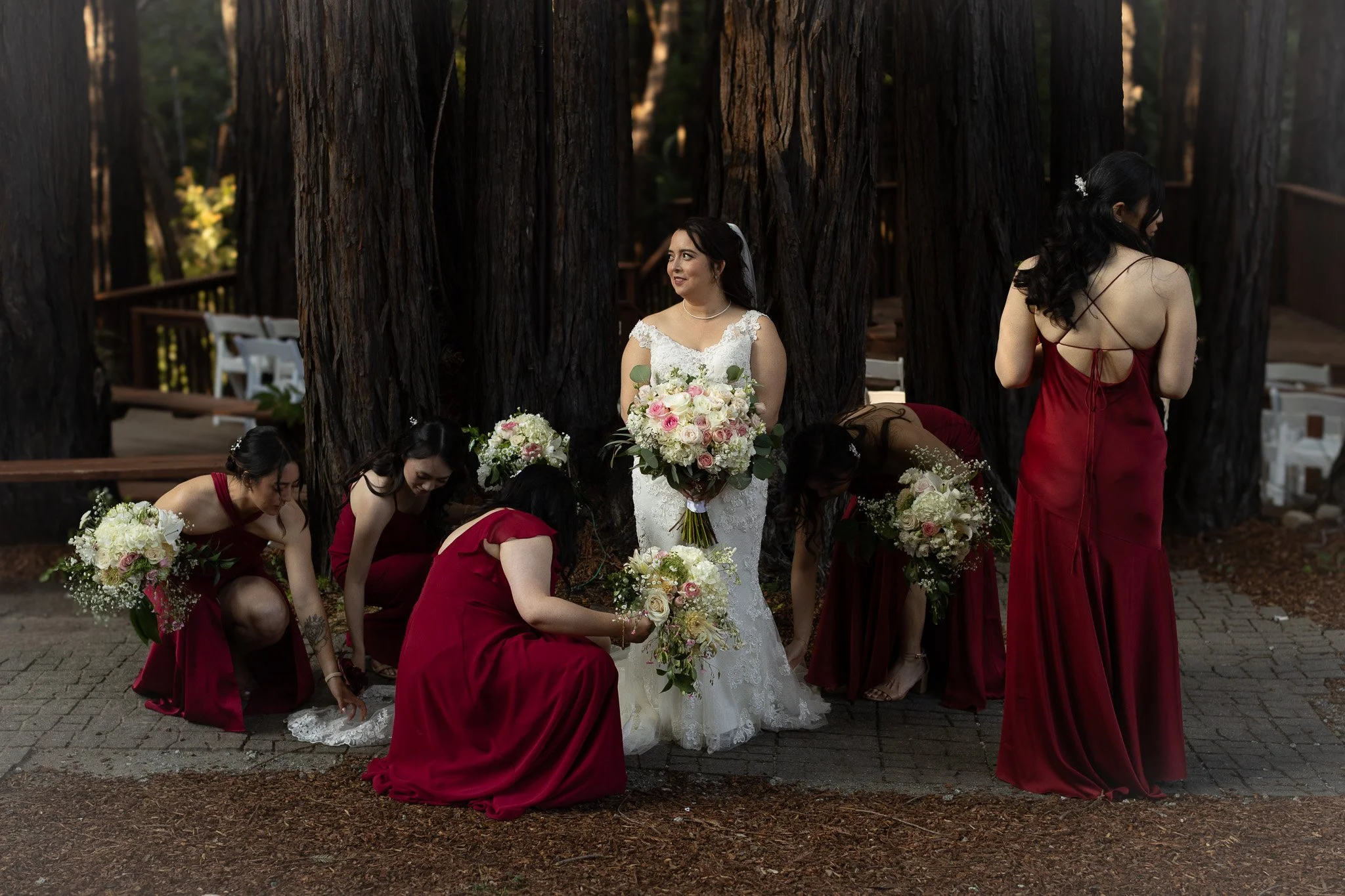 A bride in a white wedding dress with floral details, holding a bouquet of pink and white roses, standing among bridesmaids in red dresses at an outdoor pema osling redwood wedding venue.