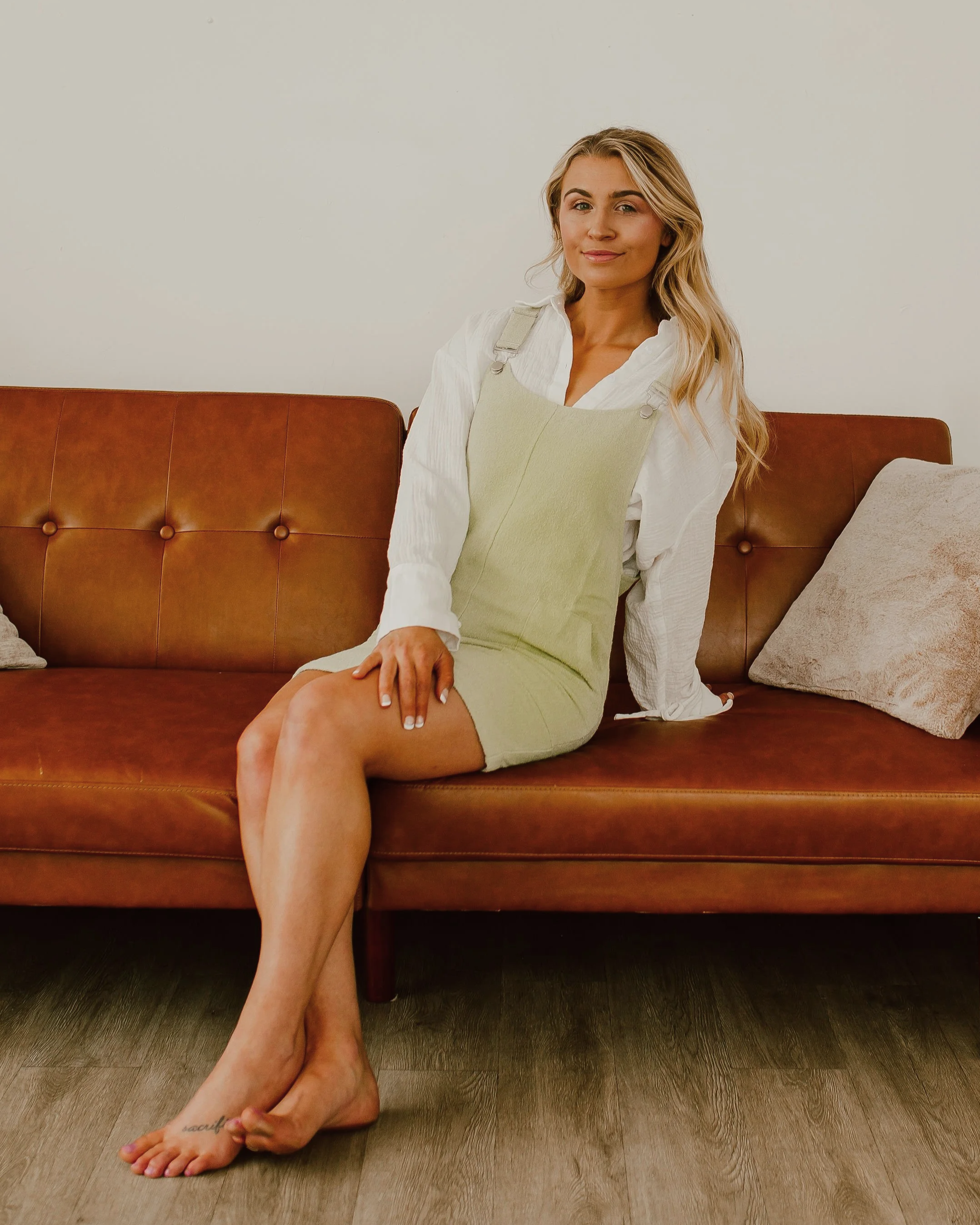 A woman with blonde hair wearing a white long sleeve shirt and a light green dress sits on a brown leather sofa with pillows, against a plain white wall.
