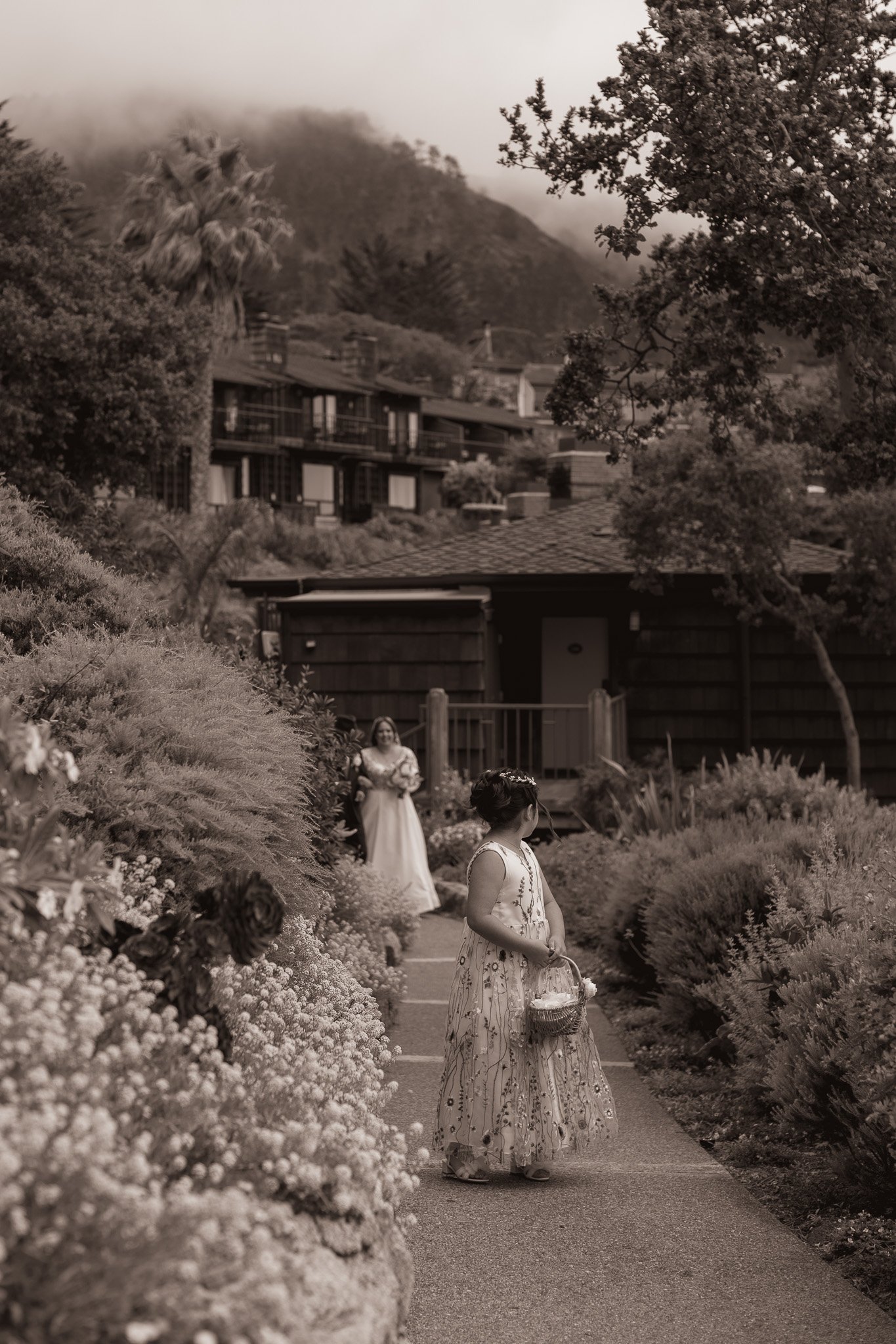 A young girl in a long floral dress holding a basket, standing on a garden path with a woman in a long dress standing in the background among blooming bushes and trees, with houses and mountains in the distance at carmel highlands hyatt hotel.