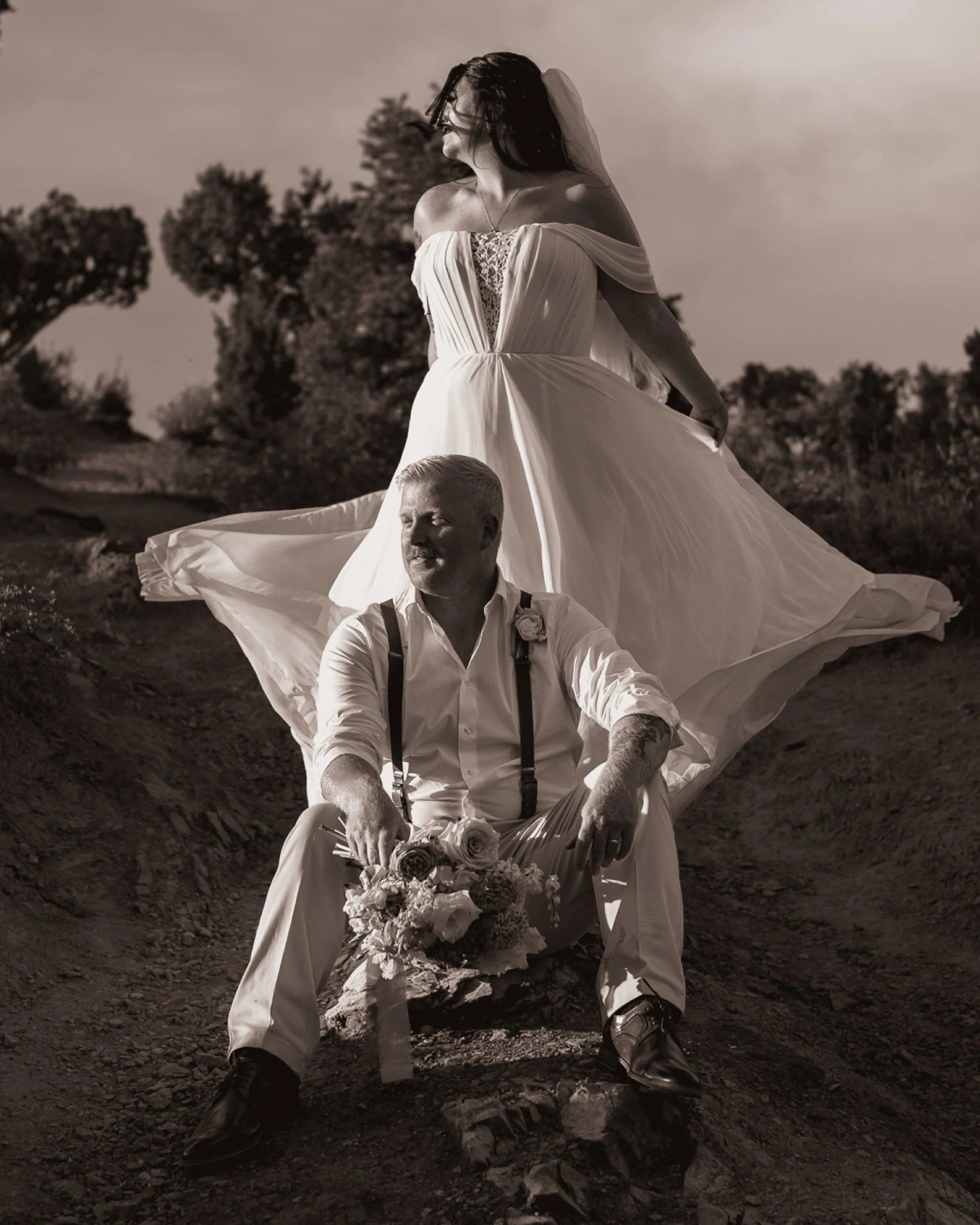 A black and white photo of a bride in a flowing wedding dress standing on a hill with trees in the background, and a man, likely her groom, sitting on the ground in front of her holding a bouquet of flowers.