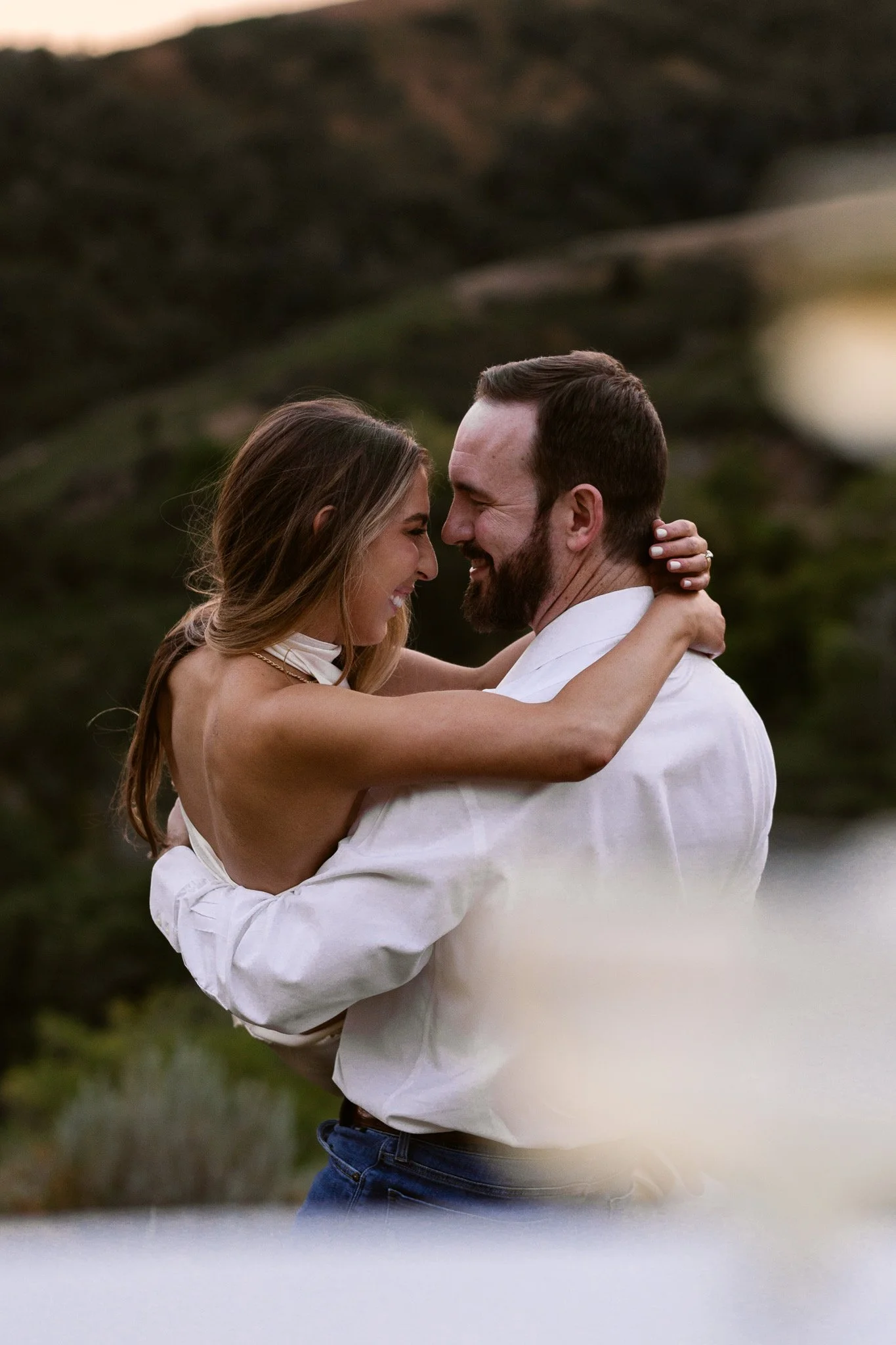A couple in casual attire embracing outdoors with hilly landscape in the background during sunset.