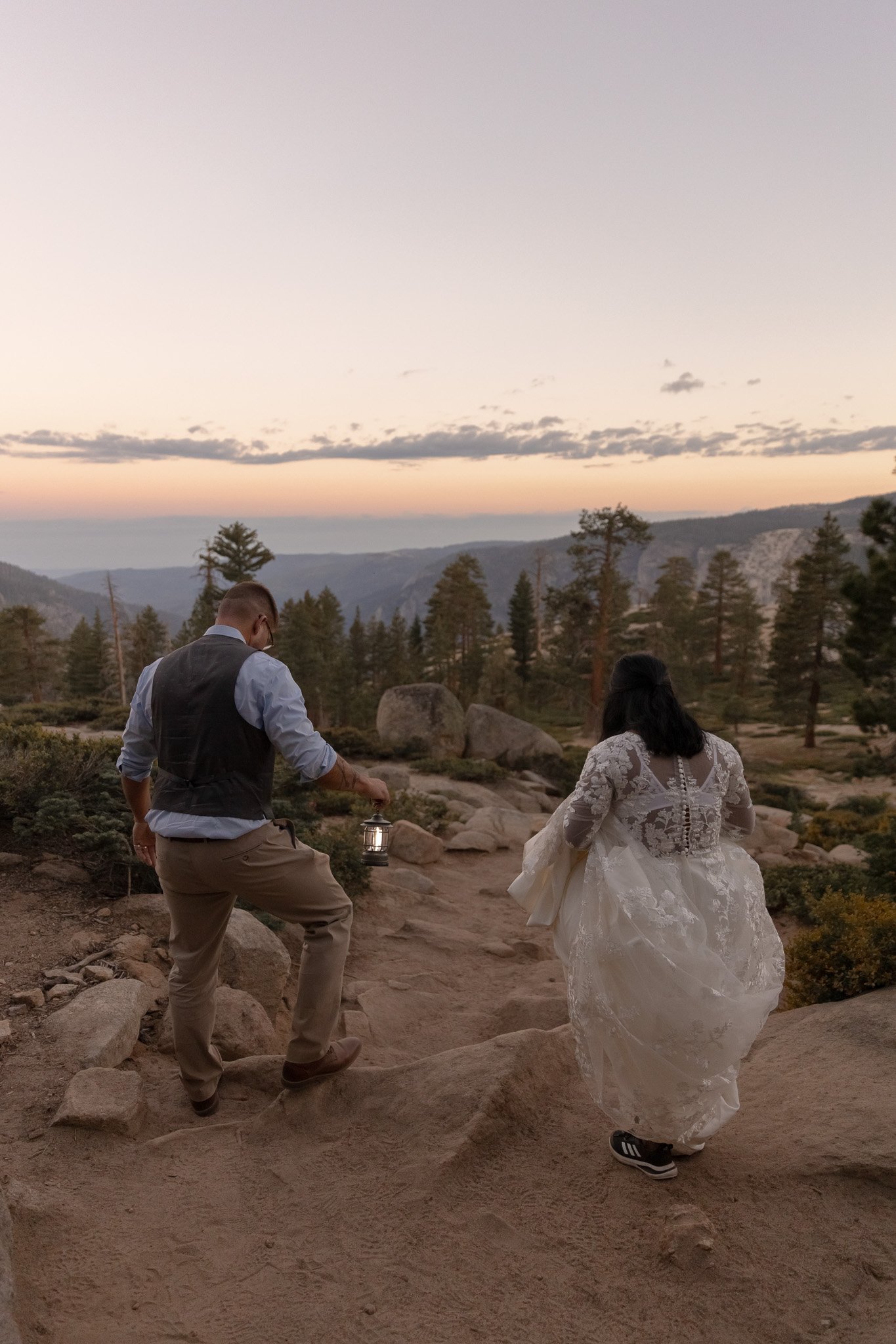 A couple walking in a rocky outdoor landscape at sunset, with the man holding a lantern and the woman wearing a wedding dress.