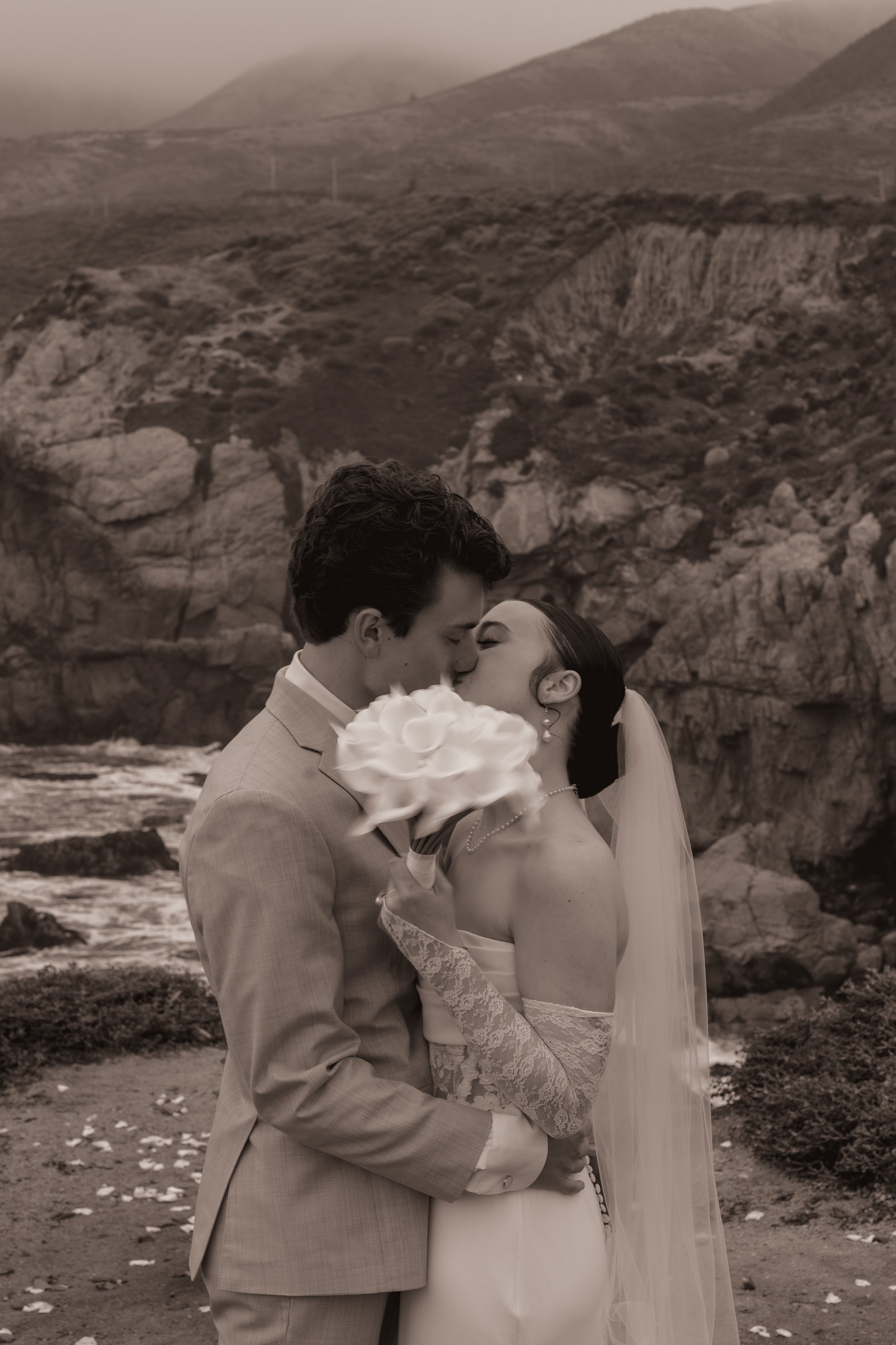 A bride and groom sharing a kiss on their wedding day outdoors, with scenic rocky hills and the sea in the background, in black and white.