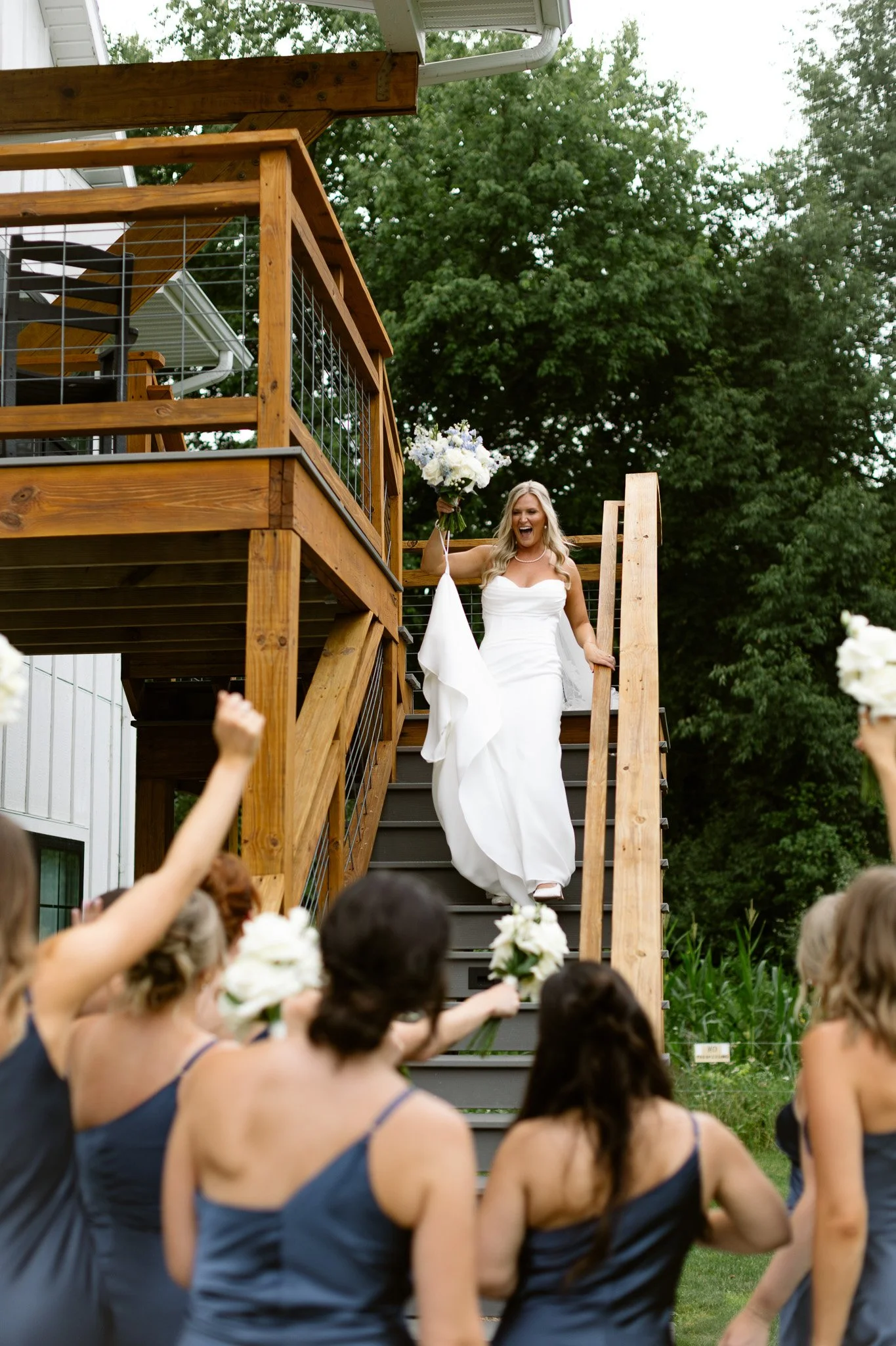 A bride in a white wedding dress holding a bouquet and celebrating with bridesmaids in navy dresses as she descends a wooden staircase during an outdoor wedding reception.