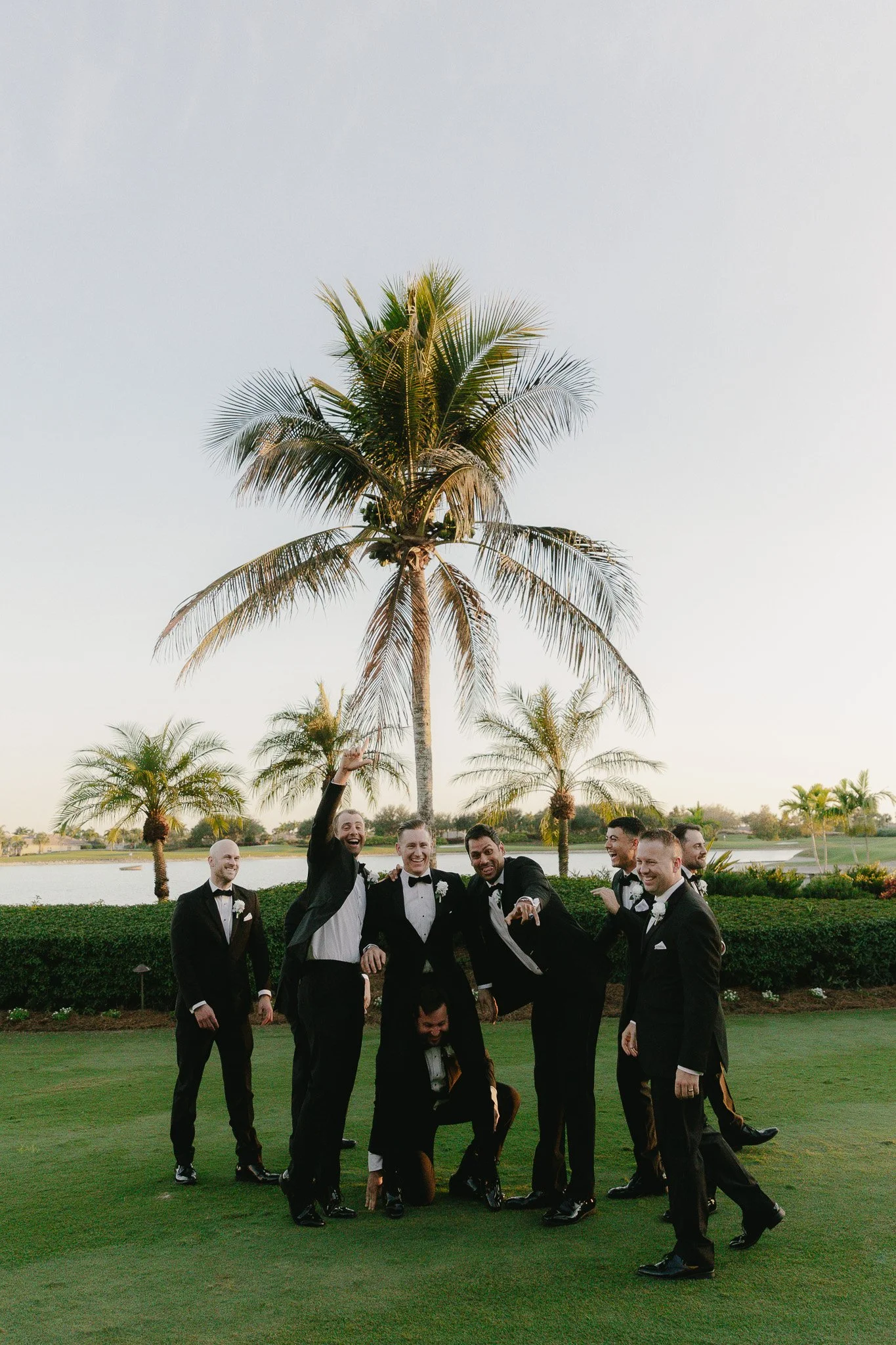 Group of men in tuxedos celebrating outdoors near a lake with palm trees in the background.