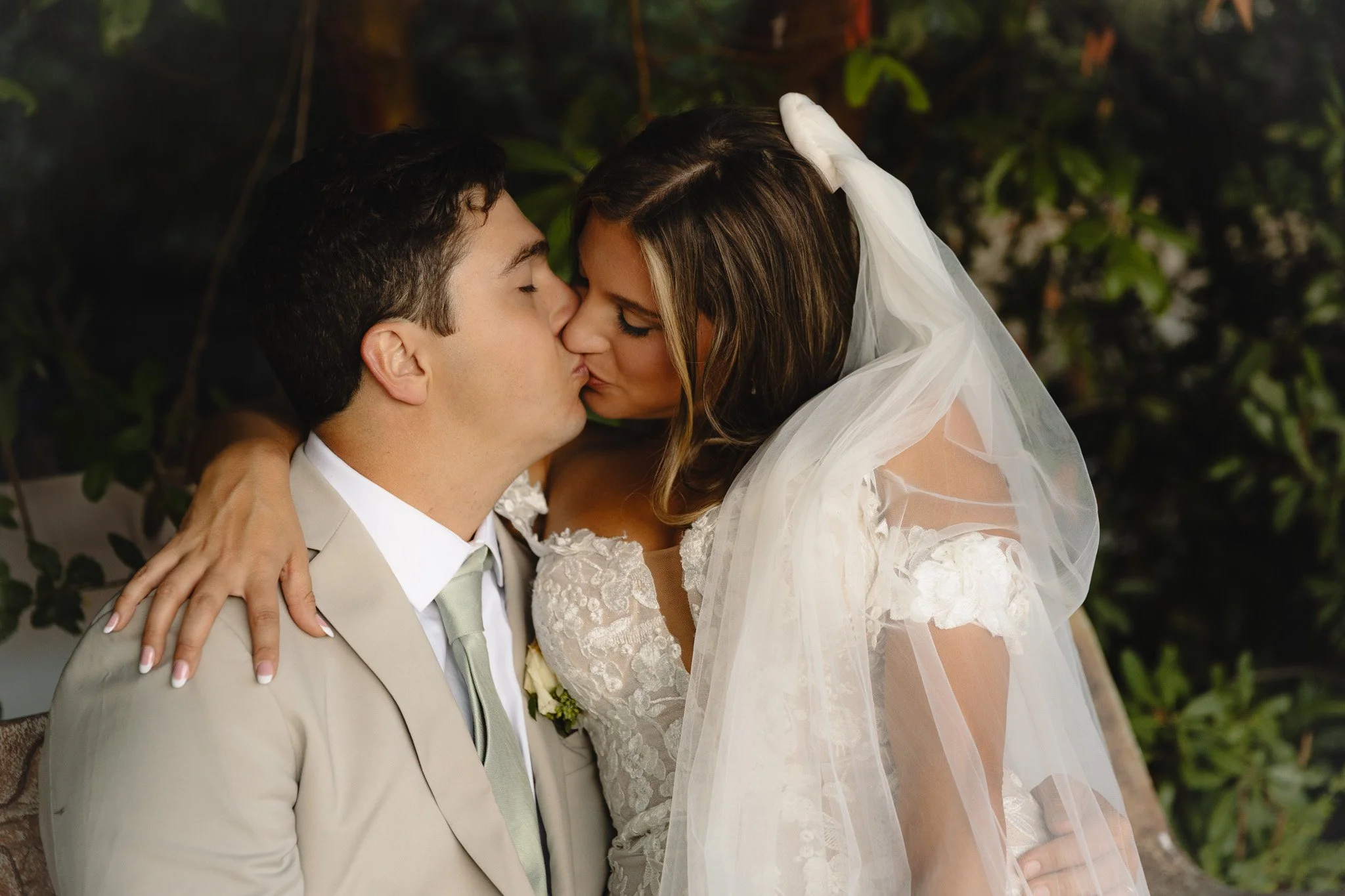 A bride and groom share a kiss on their wedding day, with the bride wearing a lace wedding gown and veil, and the groom in a beige suit with a light green tie, outdoors with green foliage in the background.