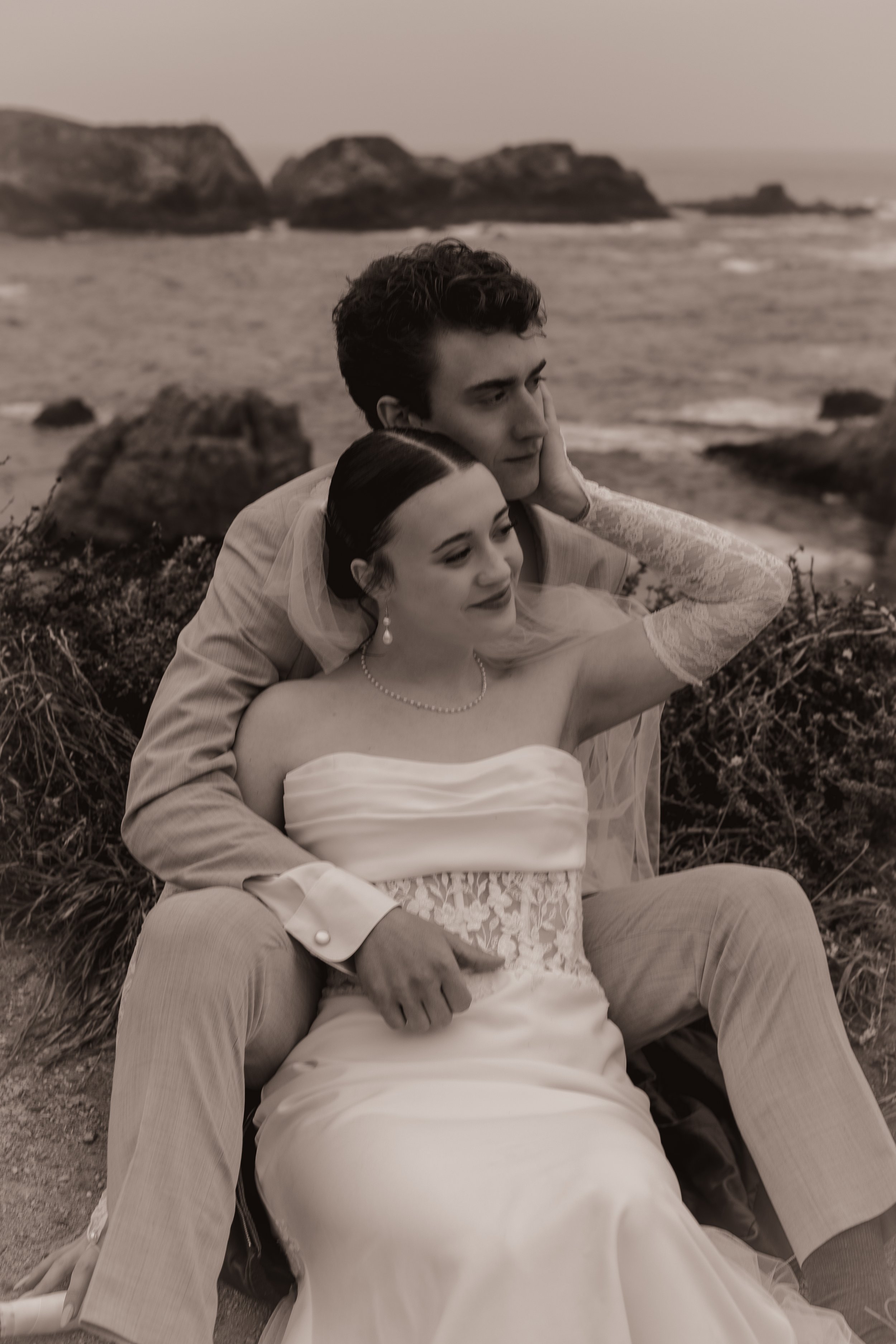 A black-and-white photo of a bride and groom sitting on the ground near the beach, with the groom holding the bride close and the ocean and rocky cliffs in the background.