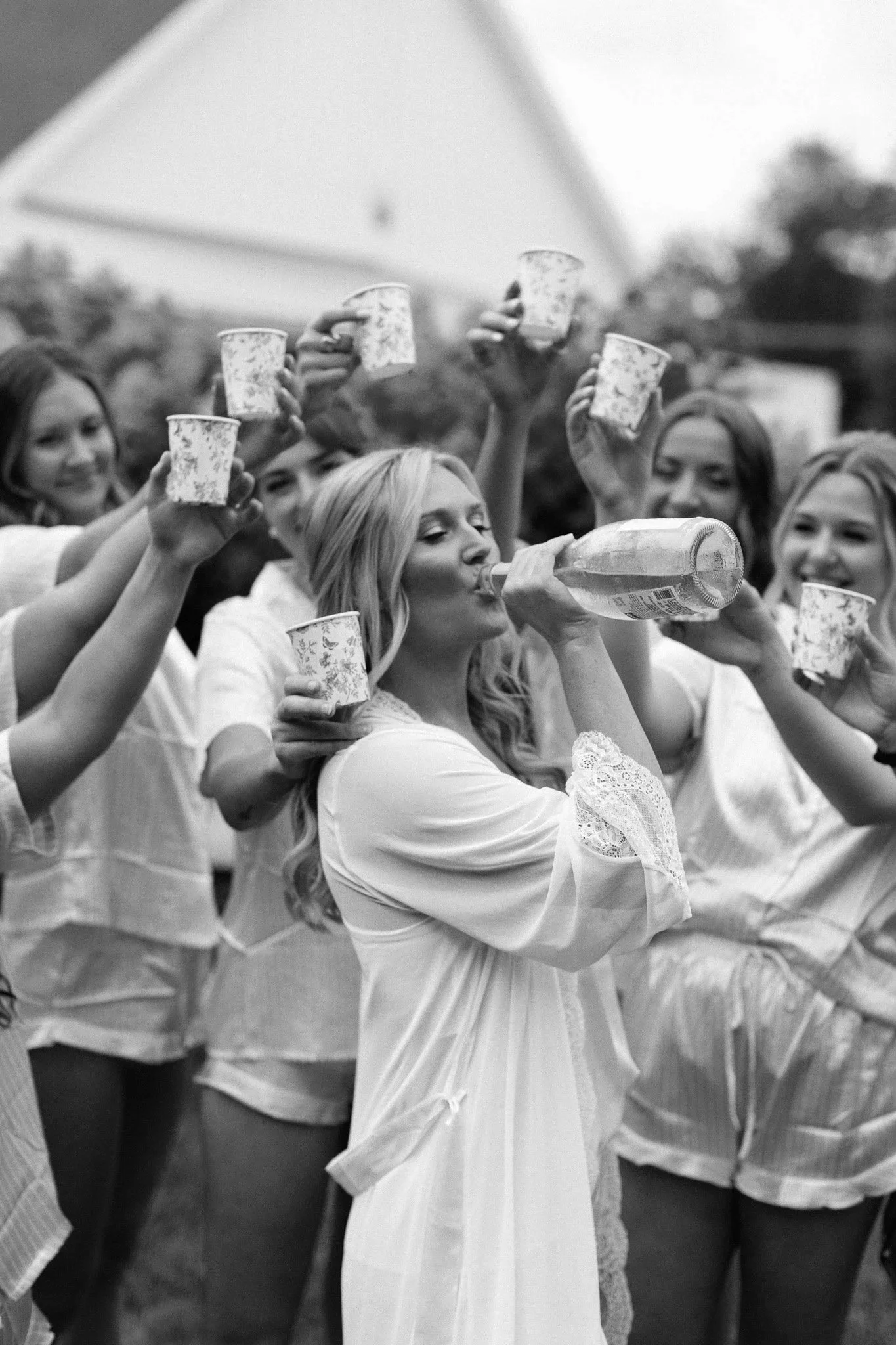 Group of women in pajamas celebrating outdoors, with one woman drinking from a bottle and others holding cups up in a toast.