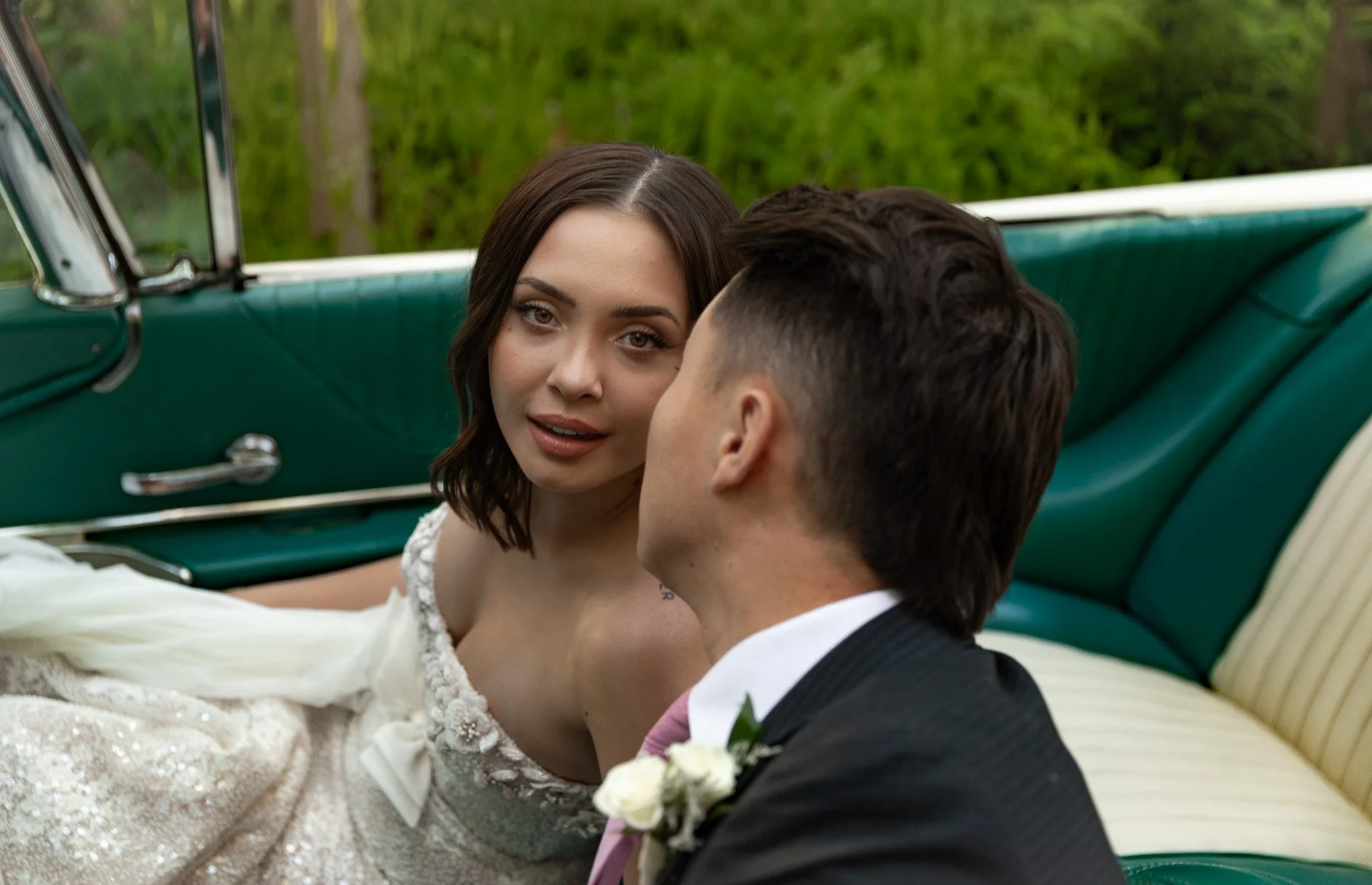 A bride and groom sitting inside a vintage car, close to each other, with the bride looking at the camera and the groom with his back turned to the camera, surrounded by green scenery at La Caille, Utah.