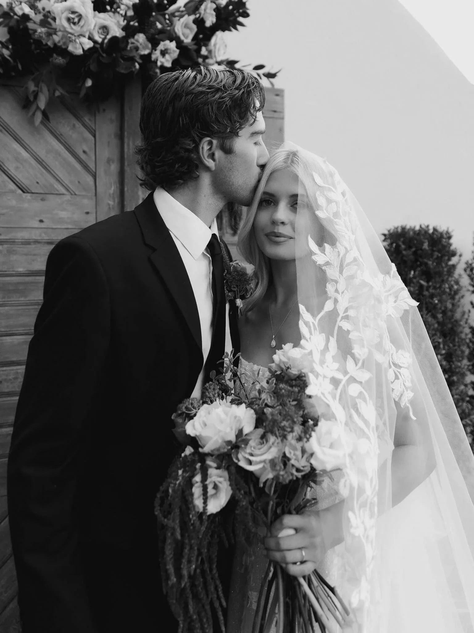 Wedding photography pricing example at Siempre Utah featuring a groom kissing bride’s forehead under floral arch with romantic veil detail