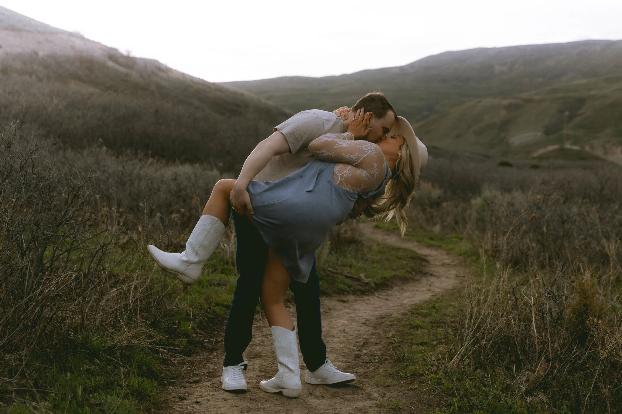 A man is lifting a woman in a light blue dress and white cowboy boots in a scenic outdoor setting with rolling hills and a dirt trail.