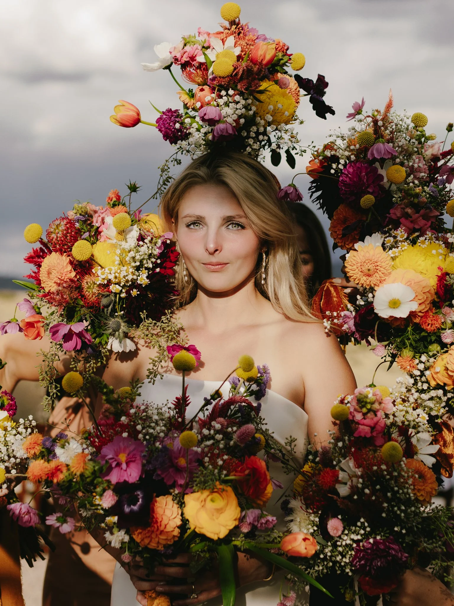A woman holding a large bouquet of colorful flowers, with more flowers around her head, against a cloudy sky background.